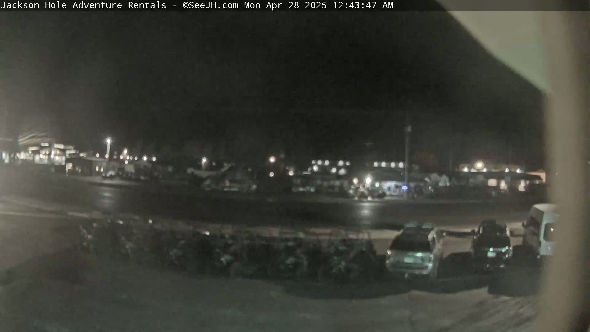 A nighttime view of a parking lot with several cars and a van, and a row of buildings in the background under a dark sky.