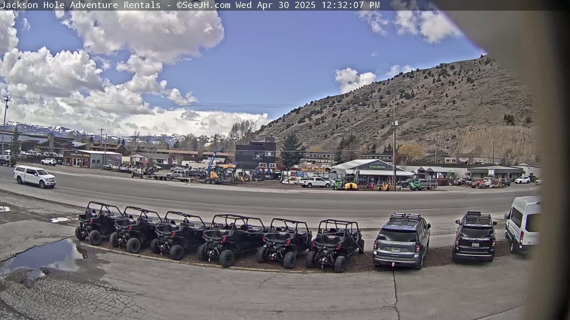 A partly cloudy day reveals a row of black ATVs parked in front of a business with several other vehicles, including SUVs and a van, near a hill and mountains in the distance.