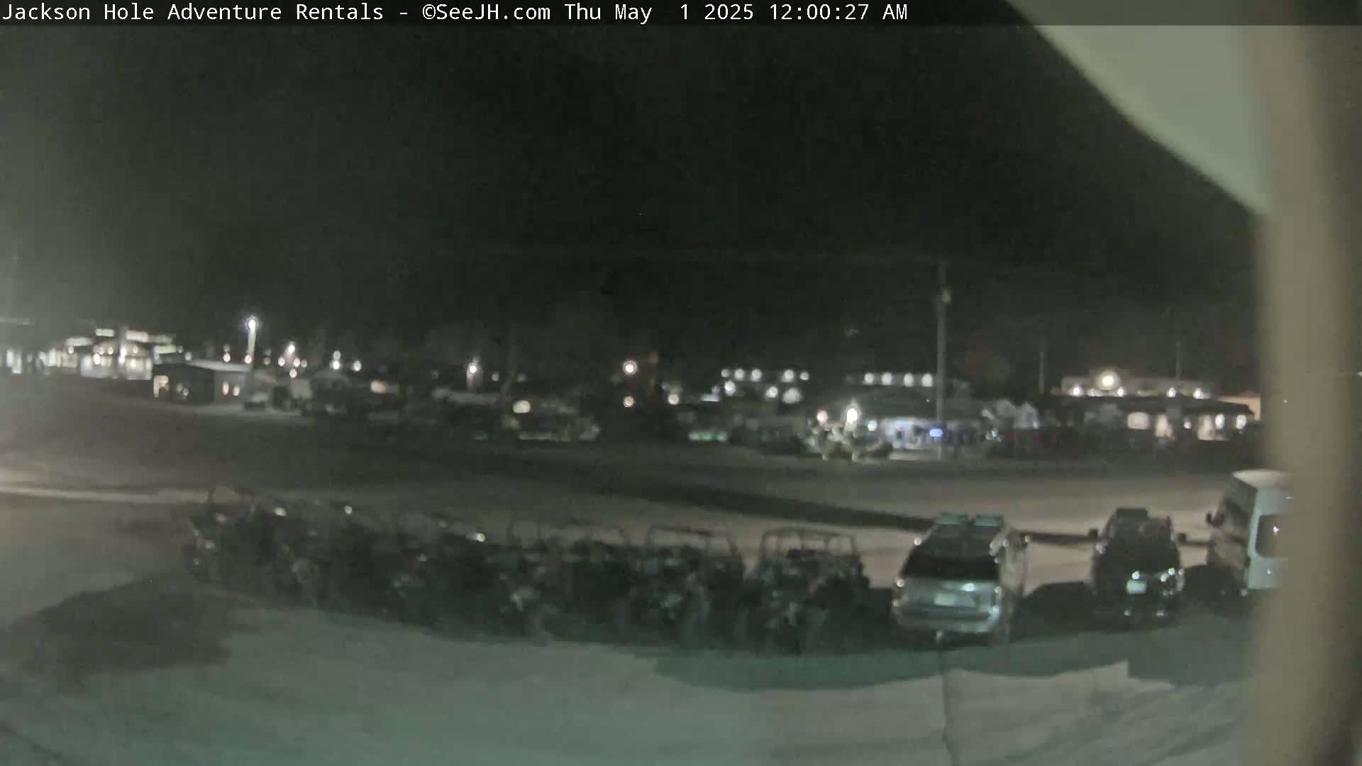 A nighttime, outdoor view shows a parking lot filled with numerous off-road vehicles and several cars, with a few buildings visible in the background under a dark sky.