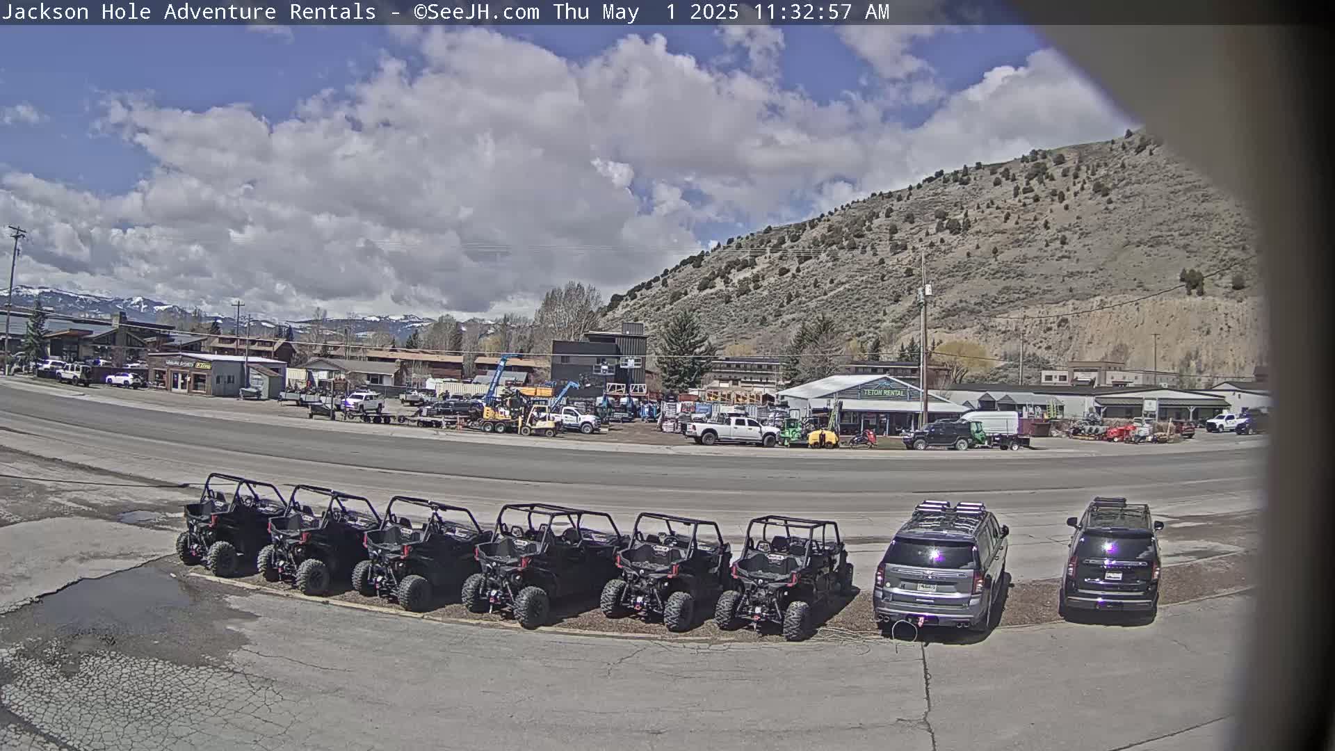 Under a partly cloudy sky, several off-road vehicles are parked in a row next to two SUVs in front of a business area with mountains in the background.