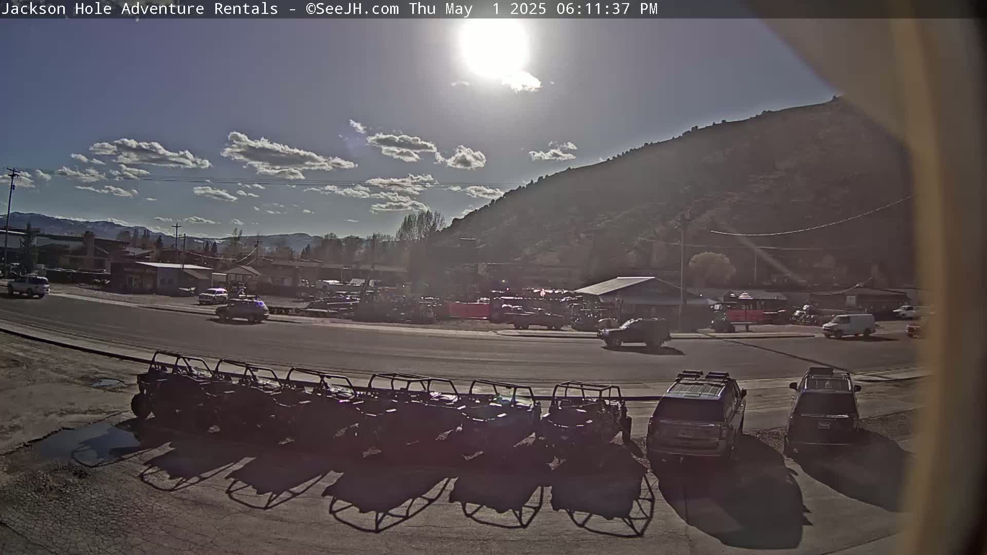 A sunny day reveals a row of all-terrain vehicles parked in a lot next to a road, with other vehicles and buildings visible in the background, nestled against a large hill.