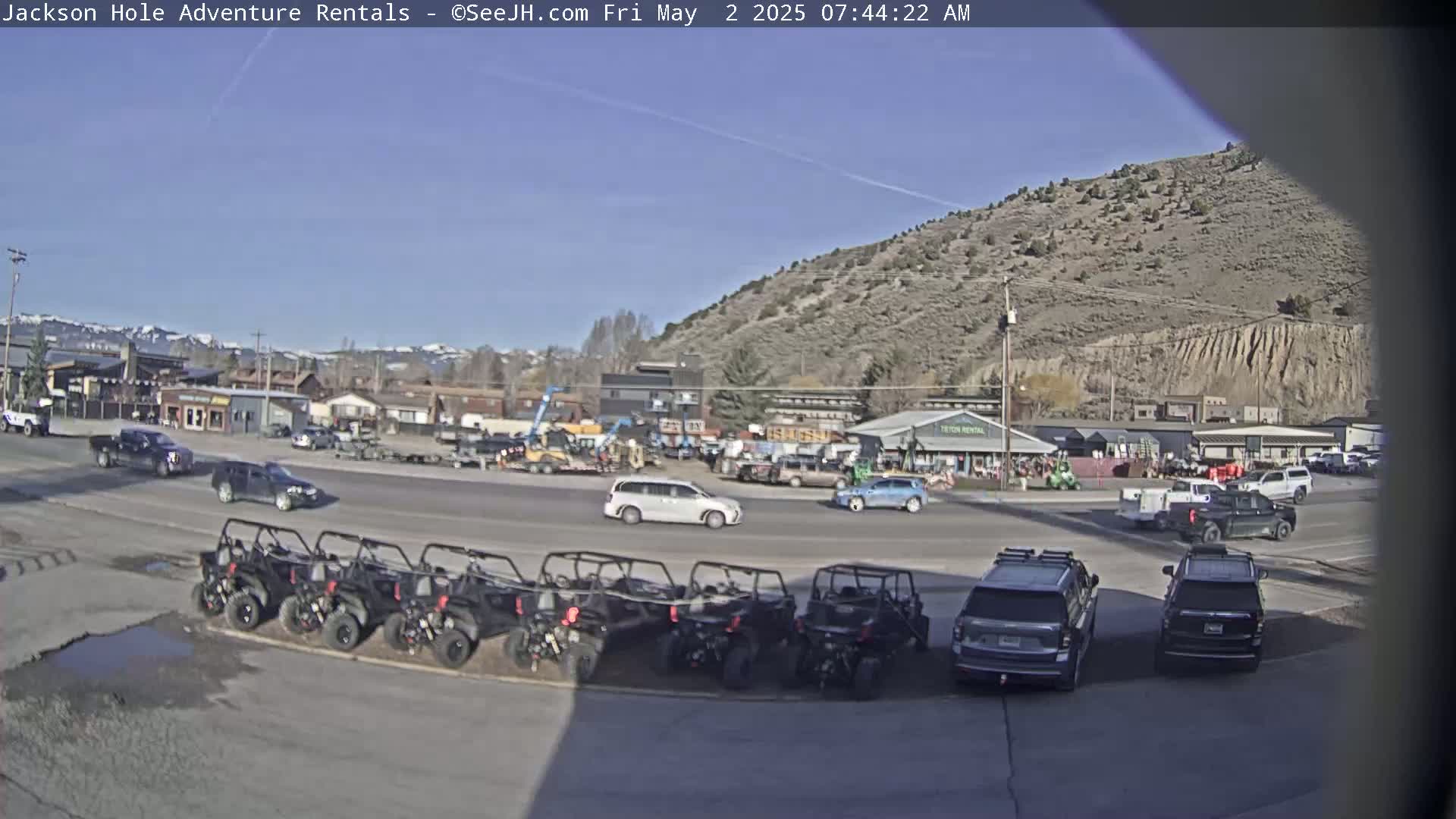 A sunny day reveals a street scene with parked cars, several all-terrain vehicles, and buildings against a backdrop of a mountainside.