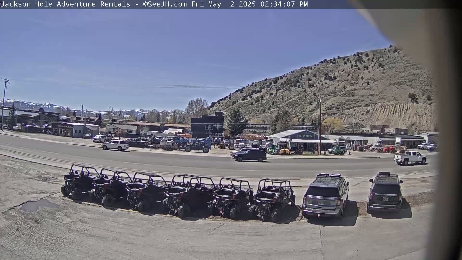 A row of all-terrain vehicles is parked in a lot next to a road, with several other vehicles and buildings in the background under a clear, sunny sky.