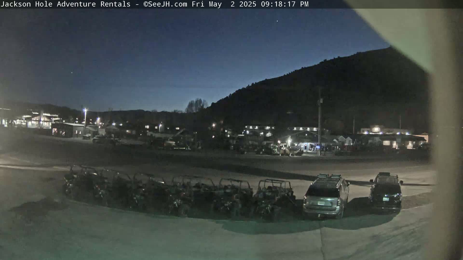 Under a dark twilight sky, a row of all-terrain vehicles is parked in a lot next to several cars in front of a collection of buildings near a dark mountain.