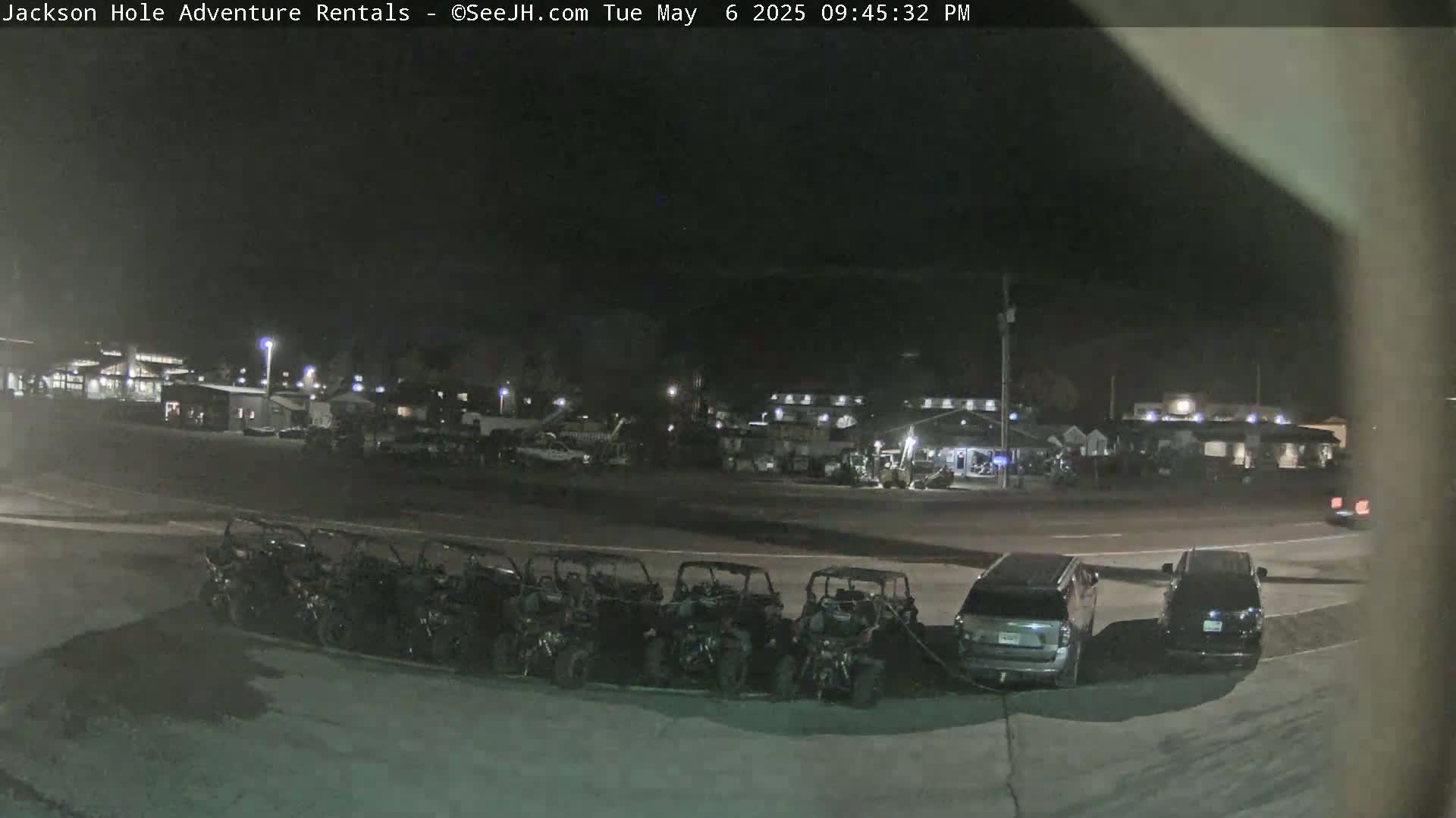 A nighttime, outdoor view shows a row of parked all-terrain vehicles next to a parking lot in front of a building complex.