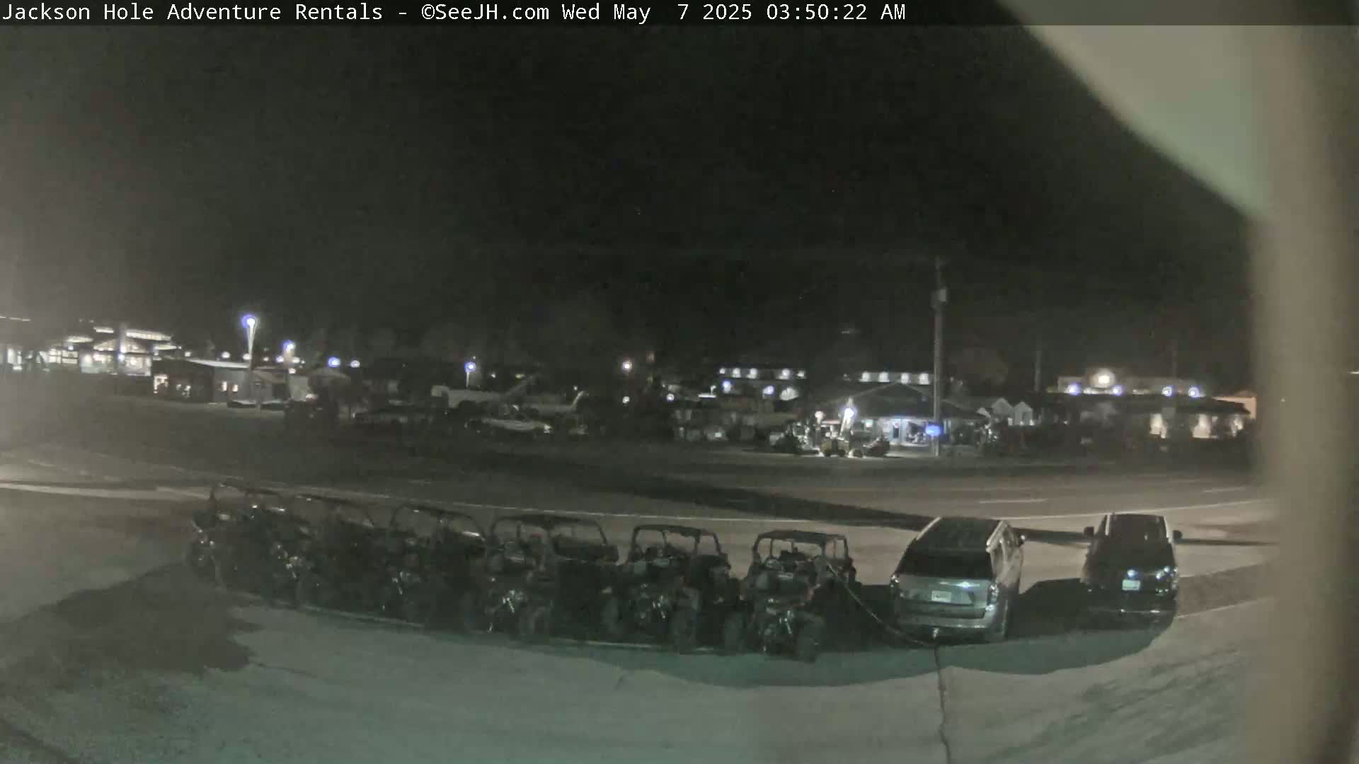 A nighttime outdoor scene shows a row of parked all-terrain vehicles in front of a parking lot with several buildings and a few cars.