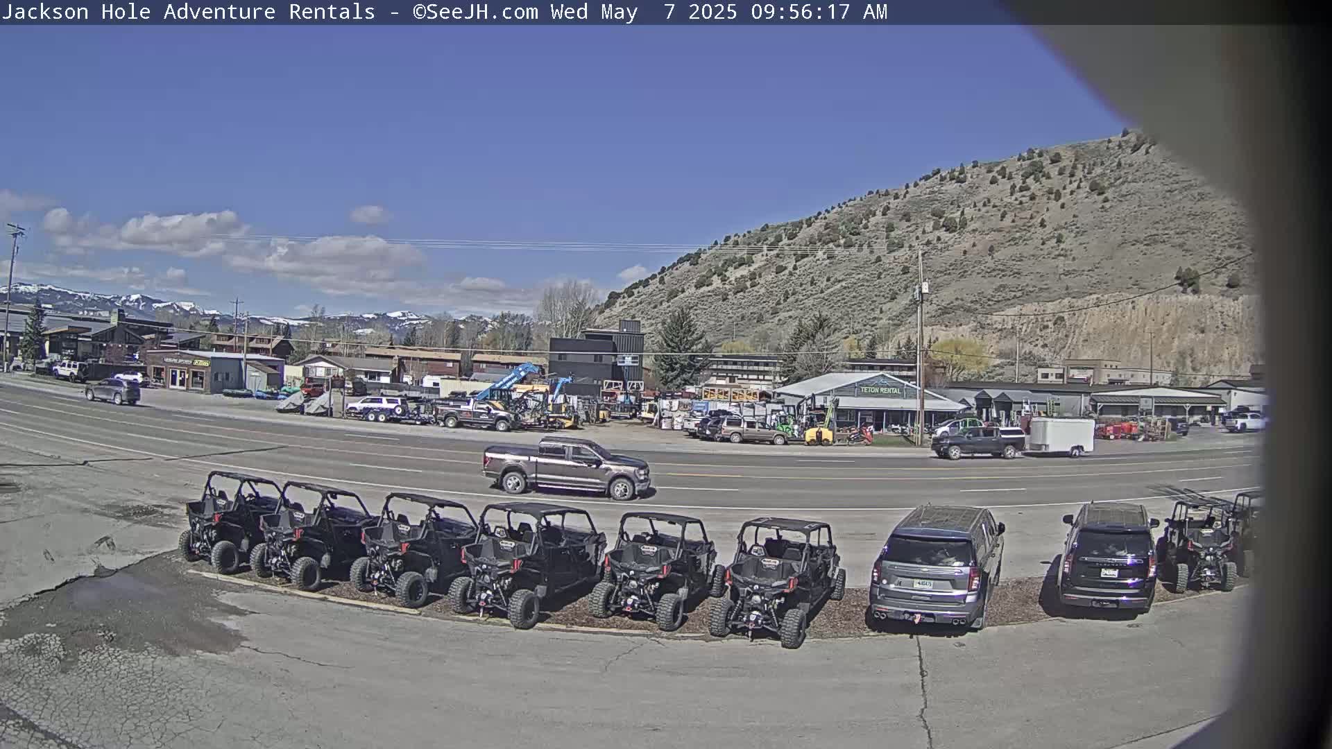 A row of off-road vehicles is parked in a lot next to a street with other vehicles and buildings, under a mostly sunny sky.