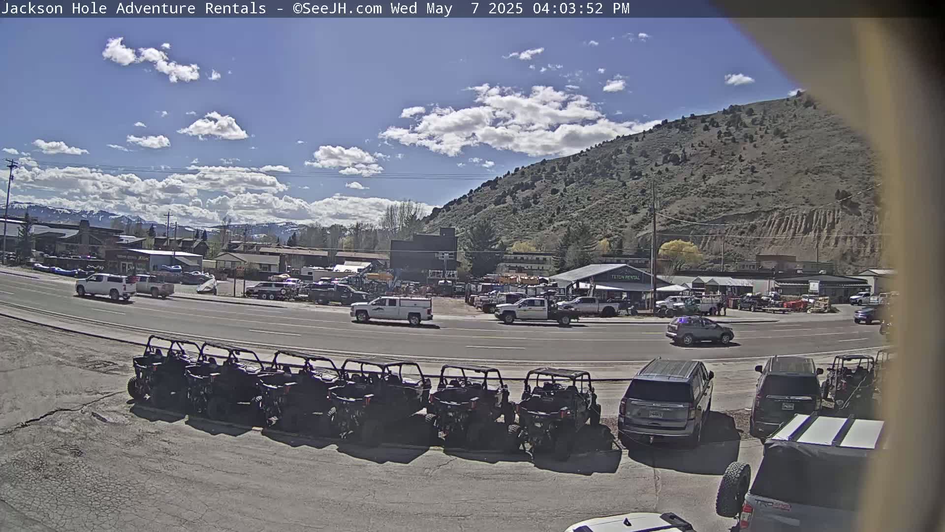 A sunny day with partly cloudy skies shows a parking lot full of off-road vehicles in front of a business and a road with several other vehicles, all situated against a backdrop of a mountainside.