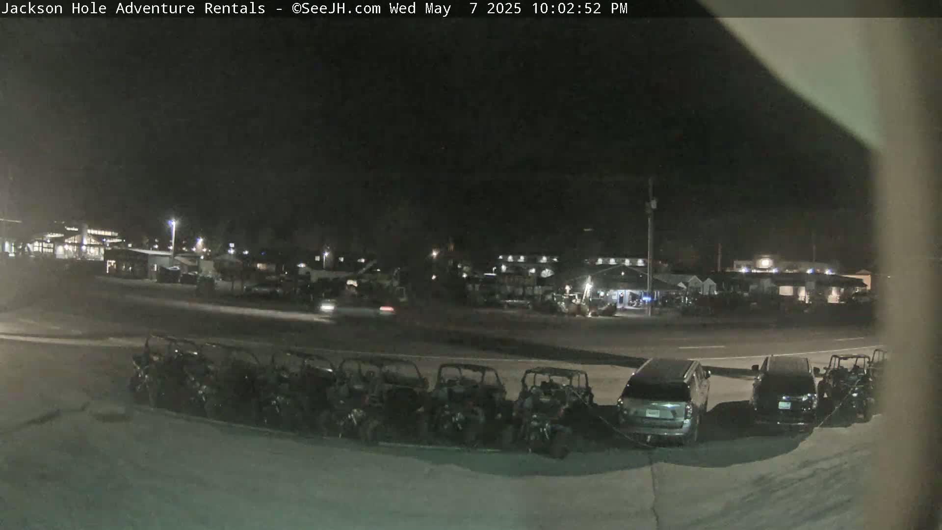 A nighttime view shows a row of parked all-terrain vehicles next to several SUVs and cars in a parking lot, with buildings visible in the background under a dark sky.