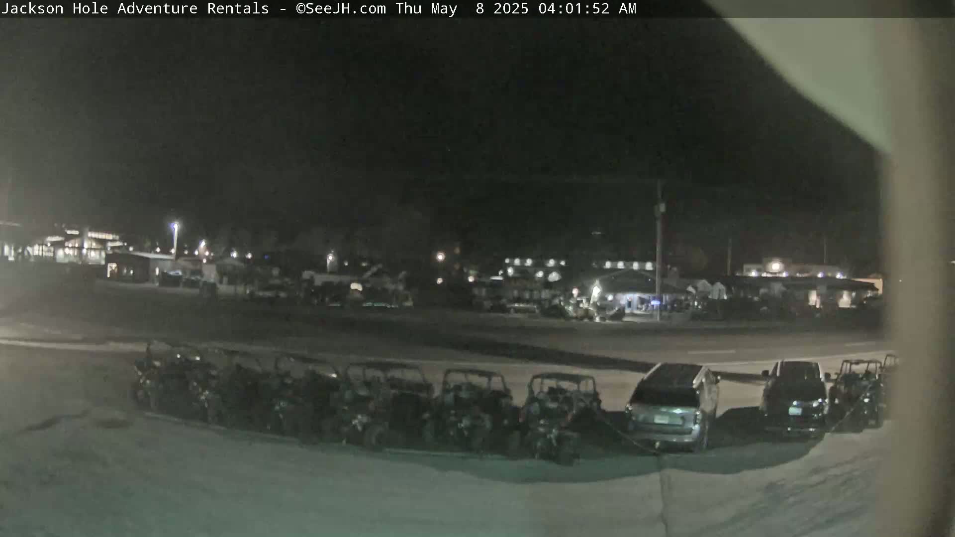 A nighttime, outdoor scene shows a row of parked all-terrain vehicles in front of a parking lot with several cars and buildings in the background.