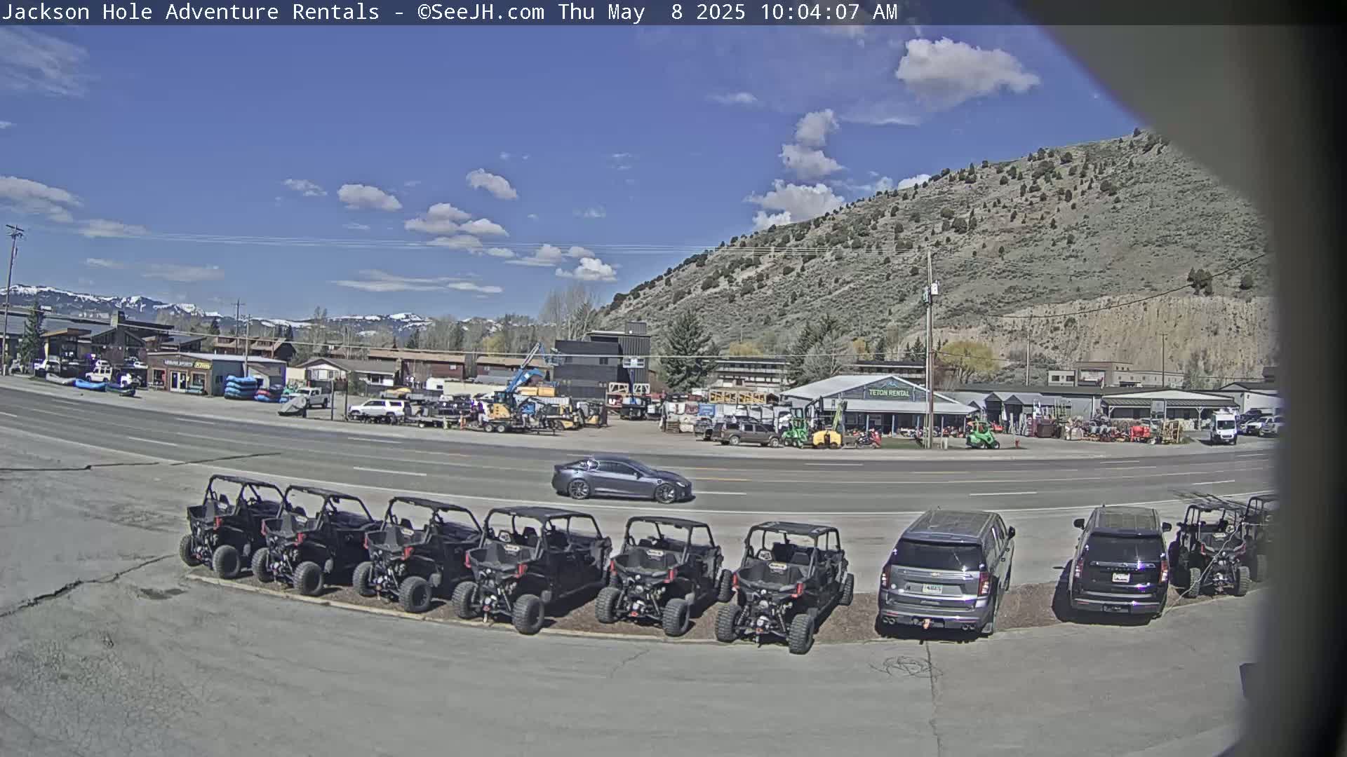A sunny day reveals a row of off-road vehicles parked in front of a business, with several cars and trucks nearby, set against a backdrop of a mountainside and a few buildings.