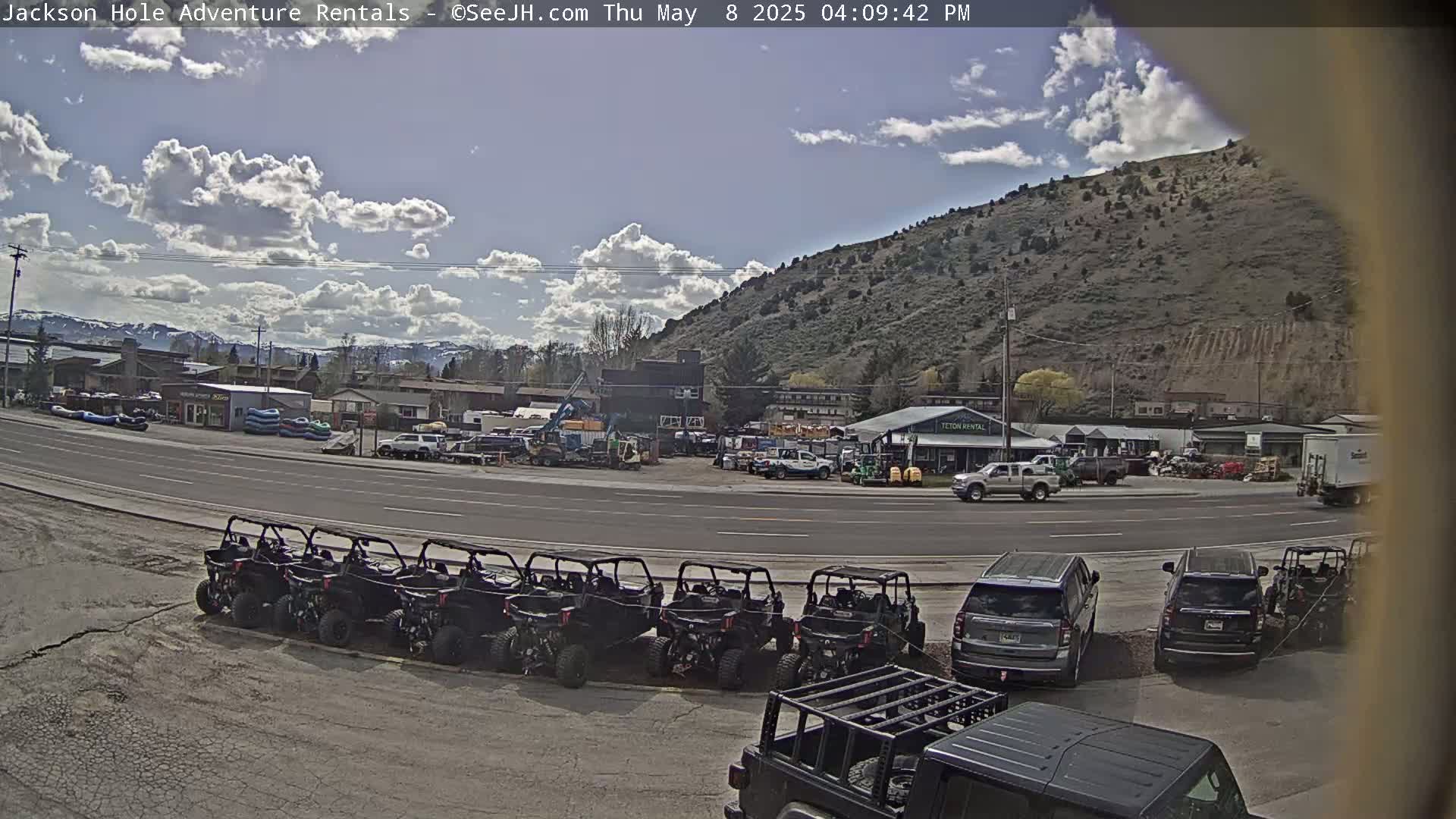 A partly cloudy day shows a street scene with numerous off-road vehicles parked in a lot next to a road with cars and trucks, and a background of buildings and a hillside.