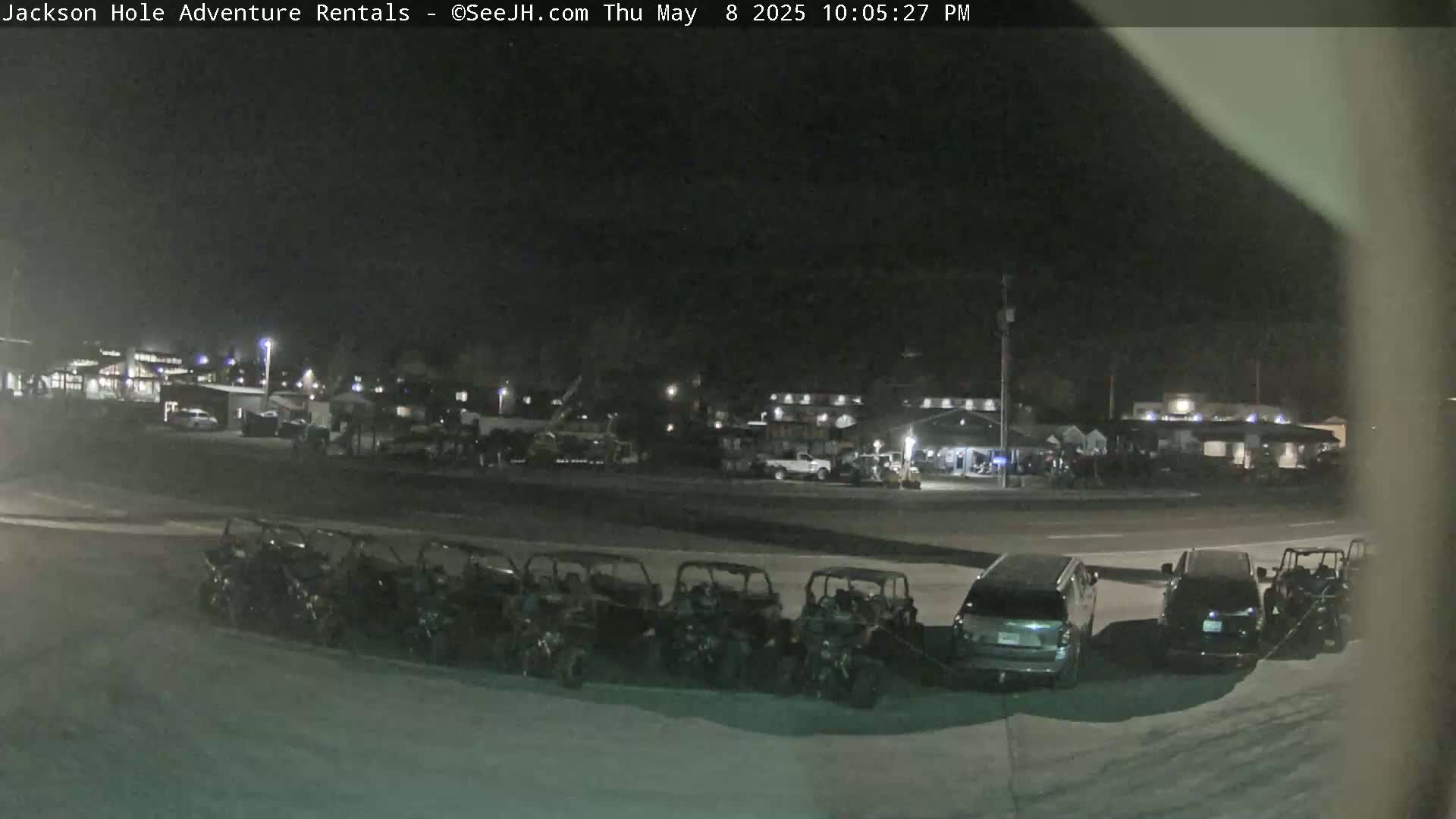 At night, several all-terrain vehicles are parked in a row near a road next to a collection of buildings under a dark sky.