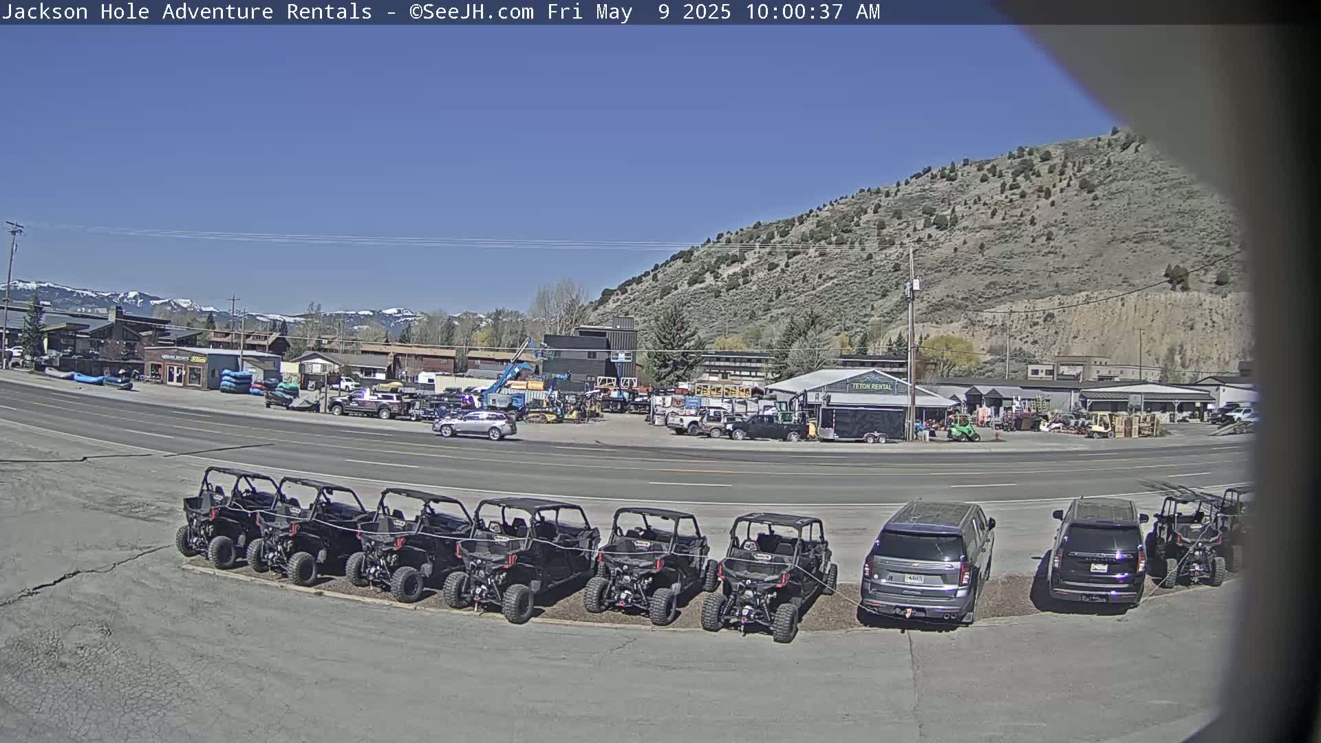 A row of off-road vehicles is parked in a lot next to a road in front of various businesses, under a clear blue sky, with mountains in the background.