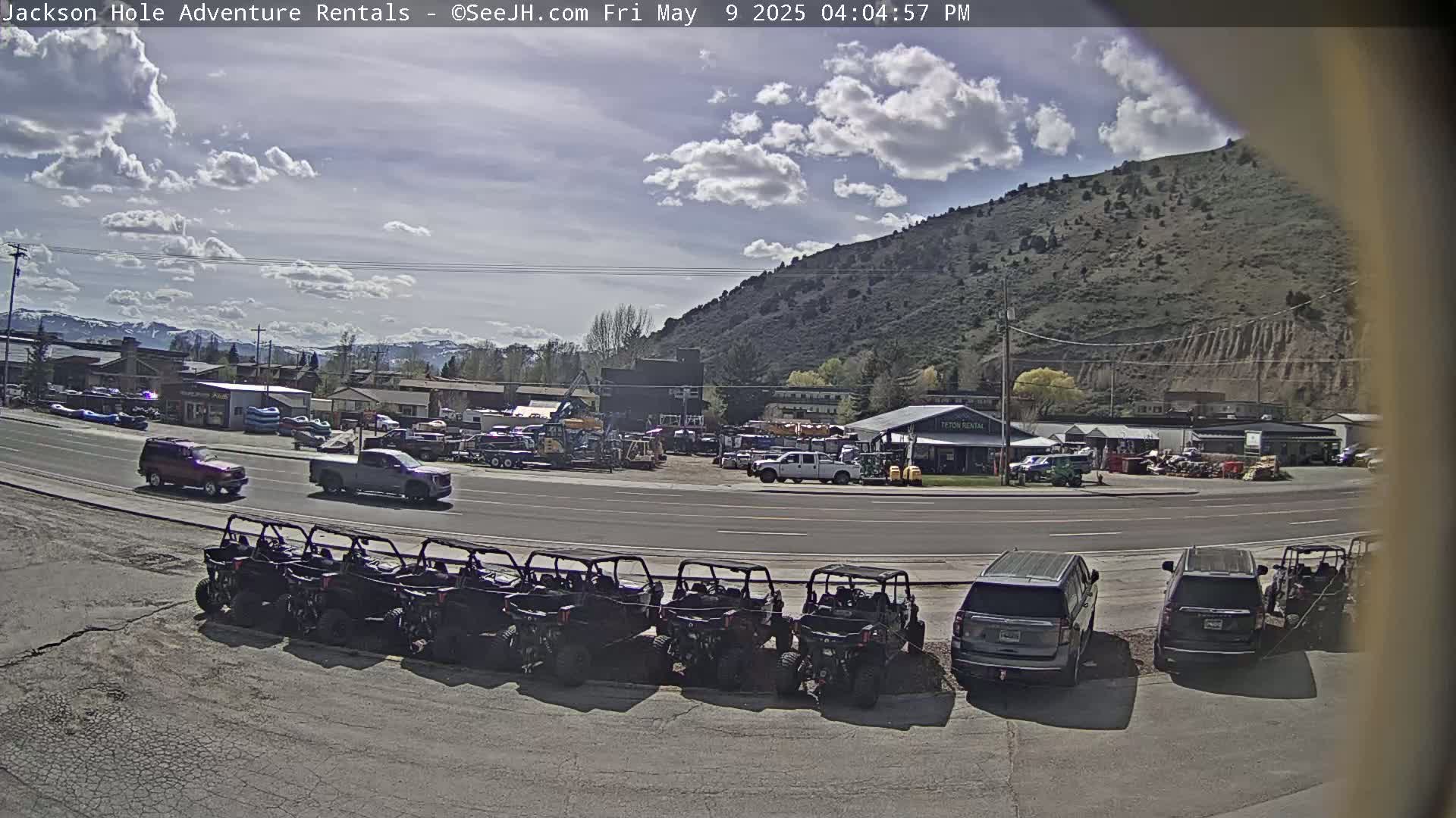 Under a partly cloudy sky, a row of off-road vehicles is parked in front of a rental business near a hill and road with cars and trucks.