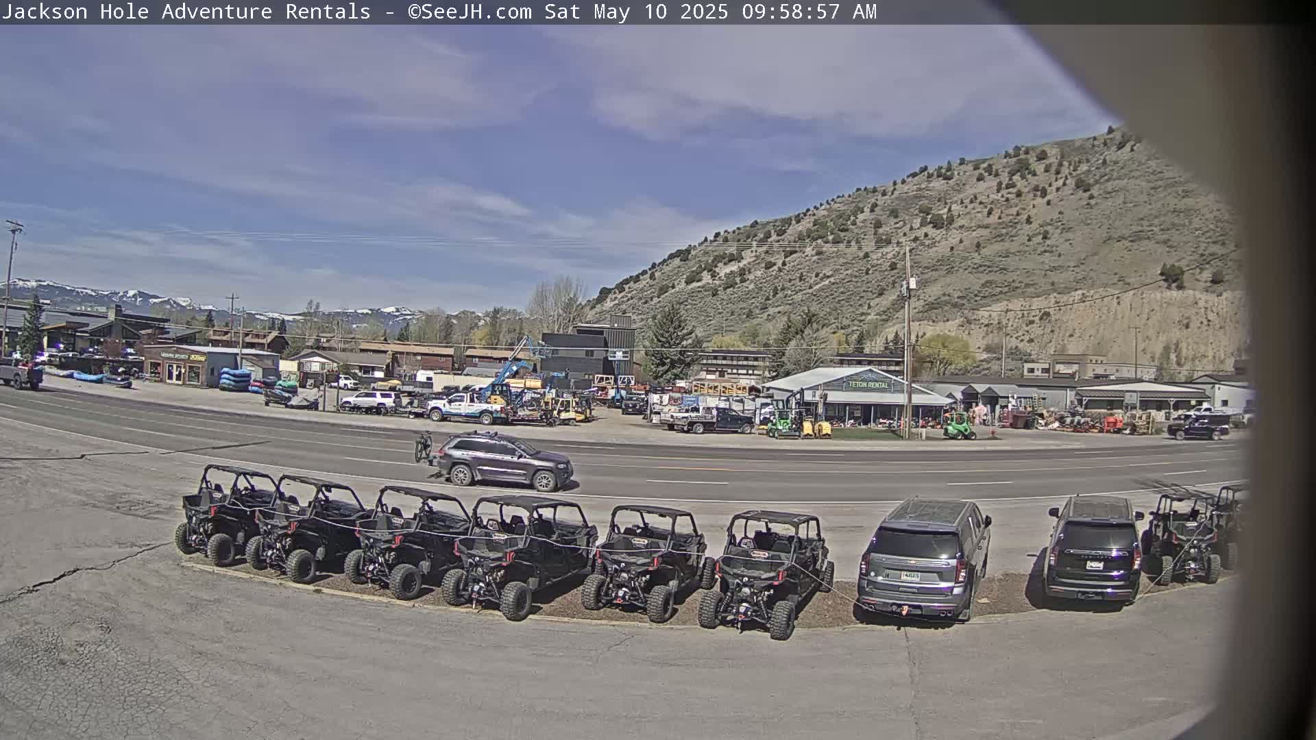 Under a mostly sunny sky, a row of off-road vehicles is parked in front of a business, across a road from other vehicles and buildings, with mountains in the background.