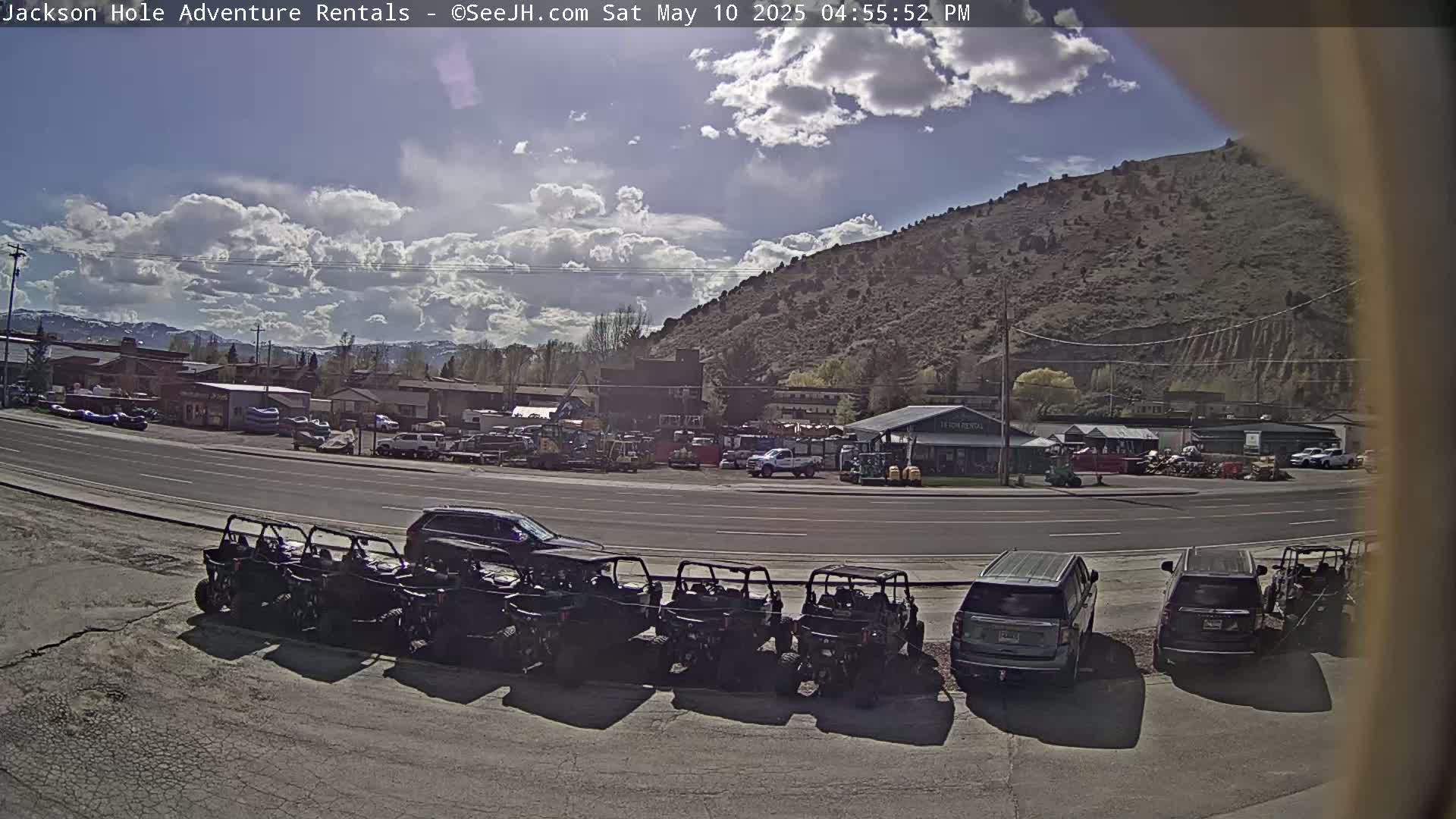 A partly sunny day shows a row of parked all-terrain vehicles in front of a building and a street with other vehicles, all set against a background of a hill and mountains.
