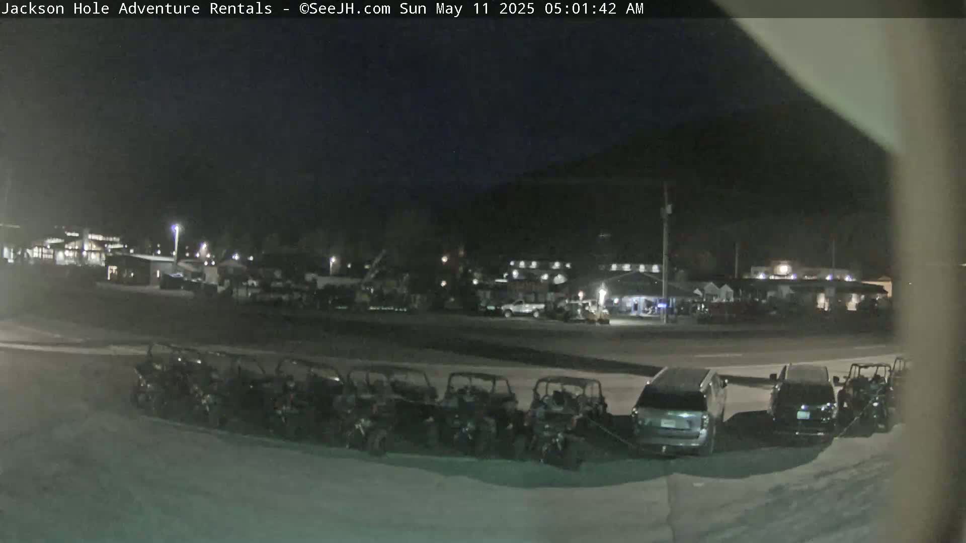 A row of all-terrain vehicles is parked at night in a lot near several buildings under a dark sky.