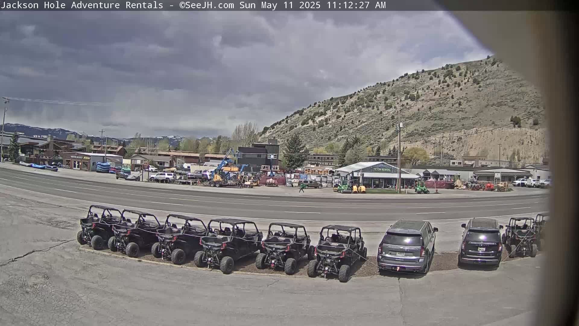 A row of off-road vehicles is parked in a lot next to a road in front of several buildings under a partly cloudy sky.