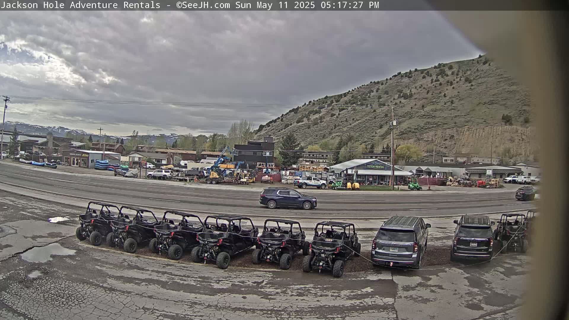 A row of black ATVs is parked in a lot next to a road in front of several buildings and a hill under a cloudy sky.