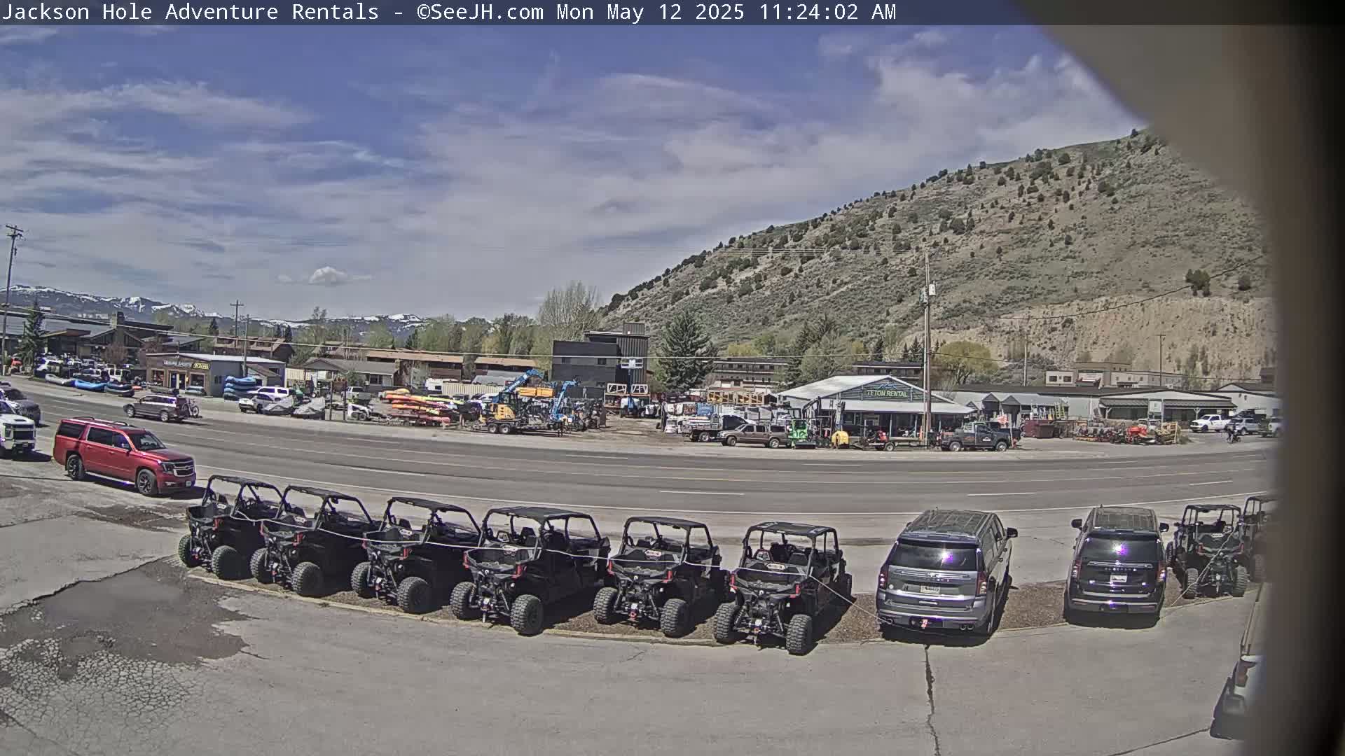 A row of all-terrain vehicles is parked in a lot next to a road in front of several businesses, with mountains in the background under a partly cloudy sky.