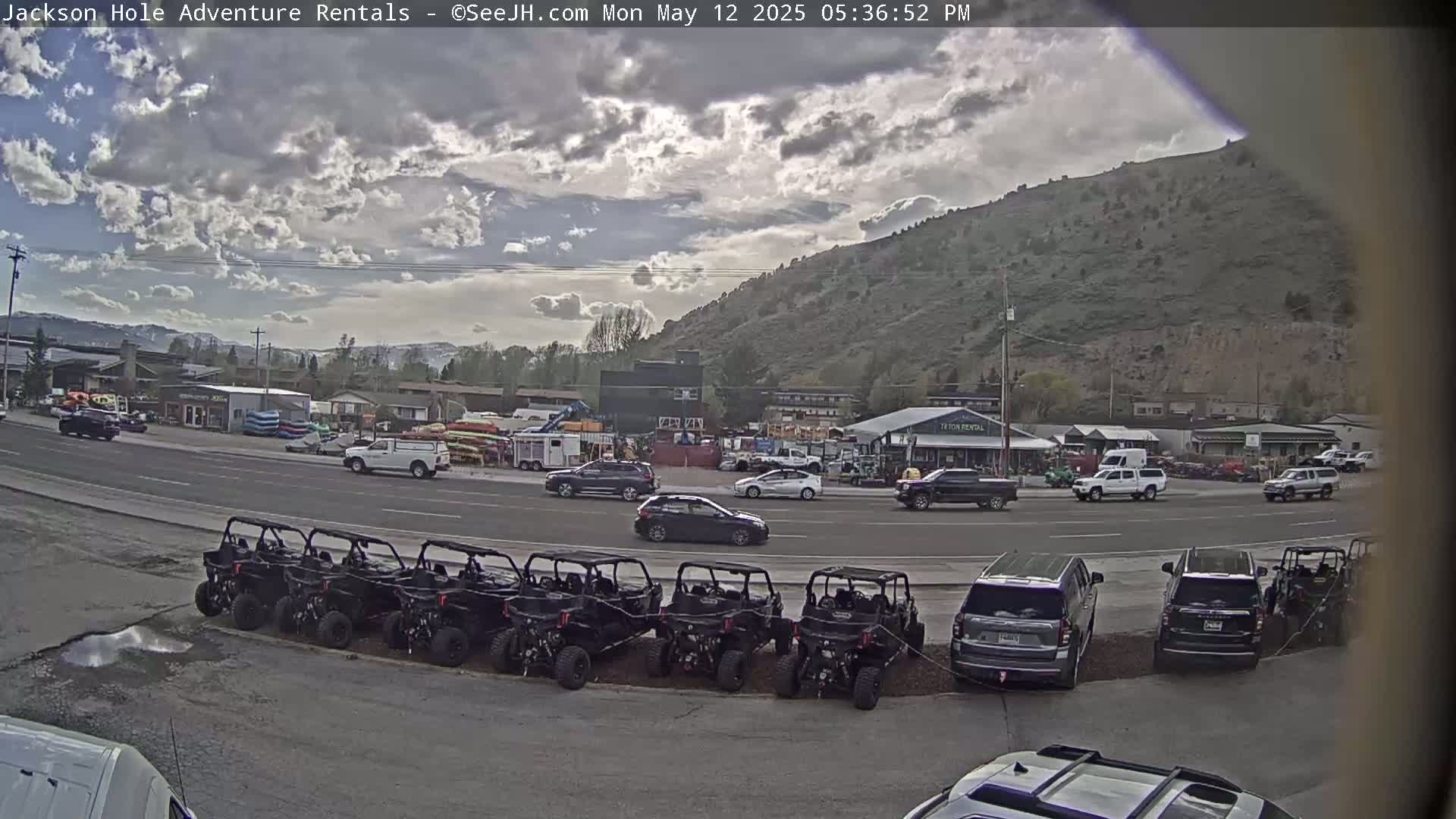A partly cloudy day reveals a row of parked all-terrain vehicles in front of a business area near a mountain, with cars and other vehicles driving on a road.
