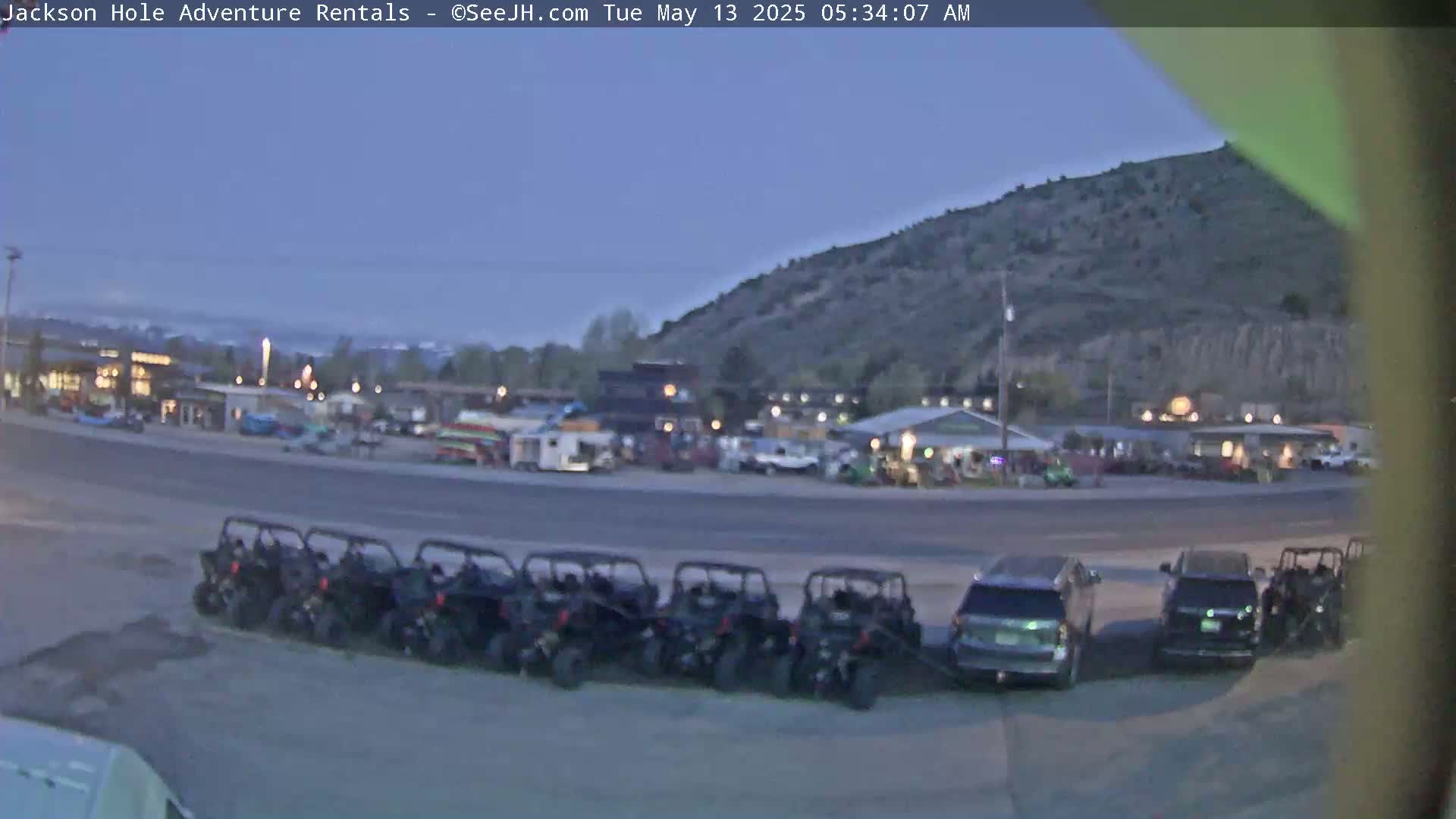 Under a twilight sky, a row of all-terrain vehicles is parked next to several SUVs in a parking lot, with buildings and a hill visible in the background.
