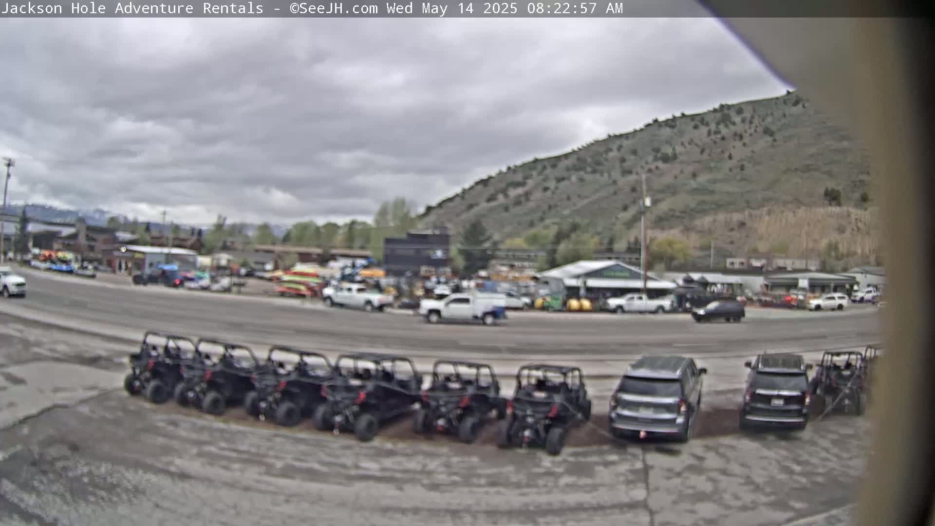 A row of all-terrain vehicles is parked in a lot next to a road, with overcast skies and a mountain in the background.