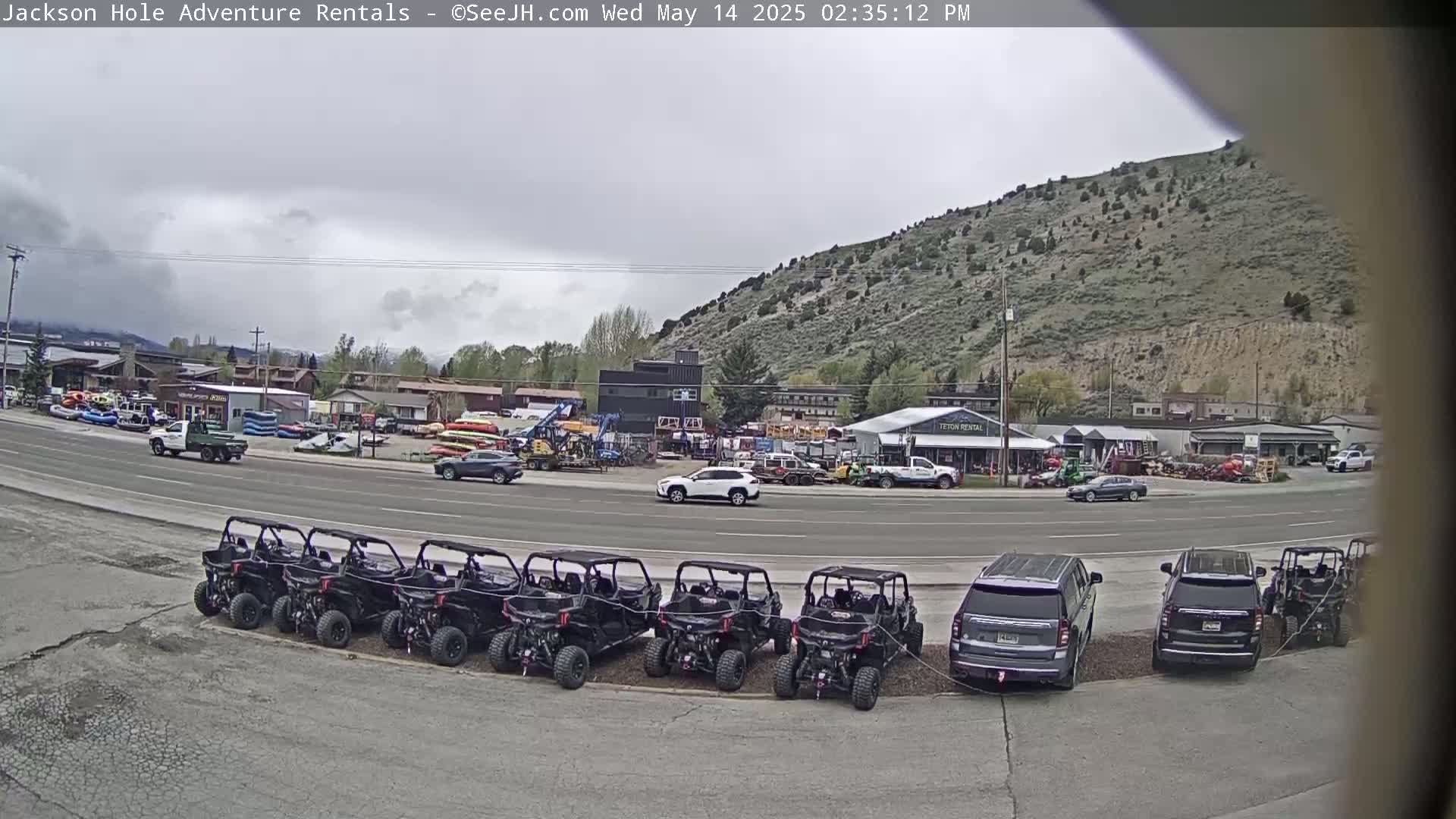 A row of black all-terrain vehicles is parked in a lot next to a street with several cars and trucks, in front of a building complex situated against a hillside under an overcast sky.
