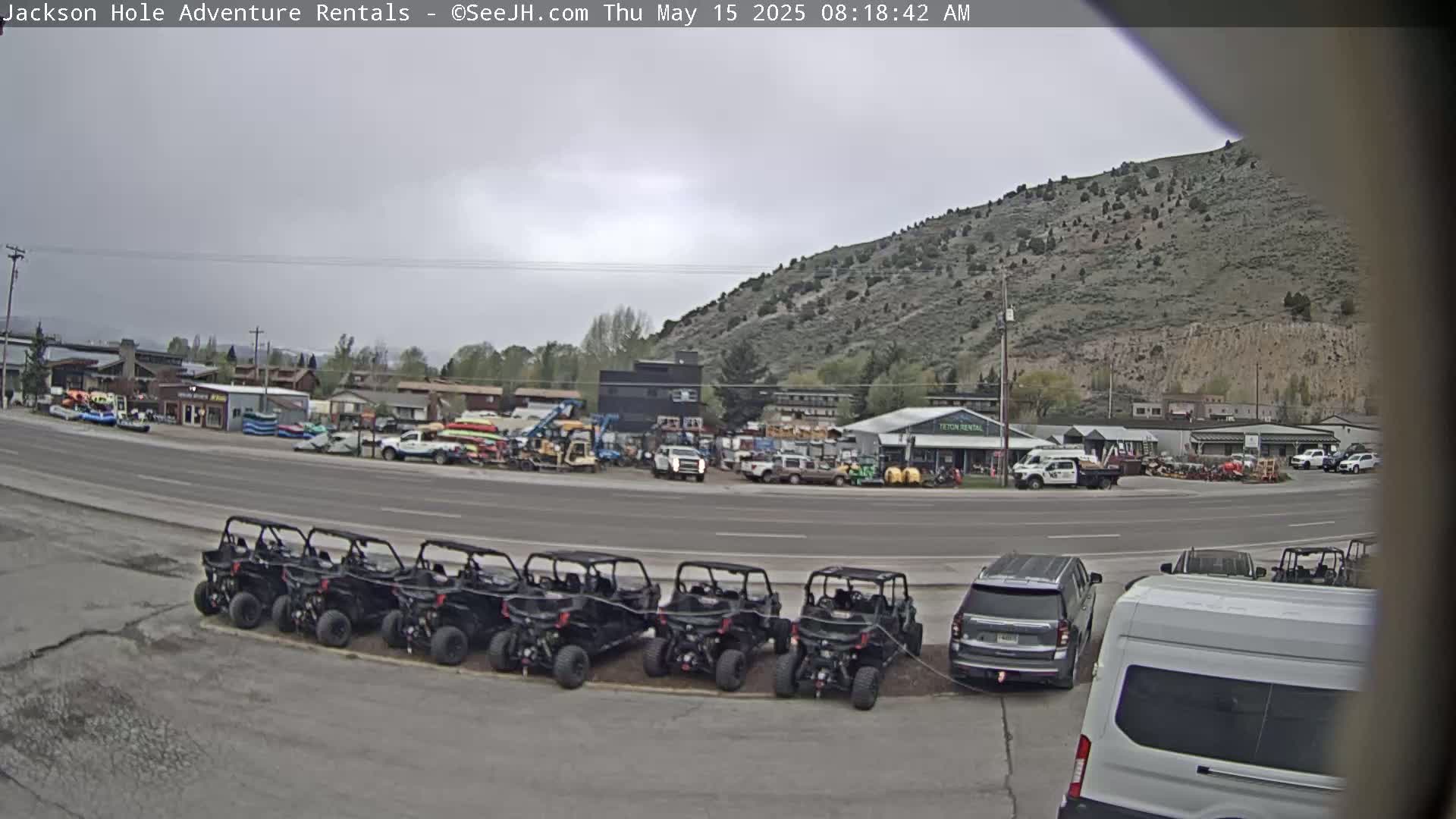 A row of all-terrain vehicles is parked in a lot next to a road, with businesses and a hill visible in the background under an overcast sky.