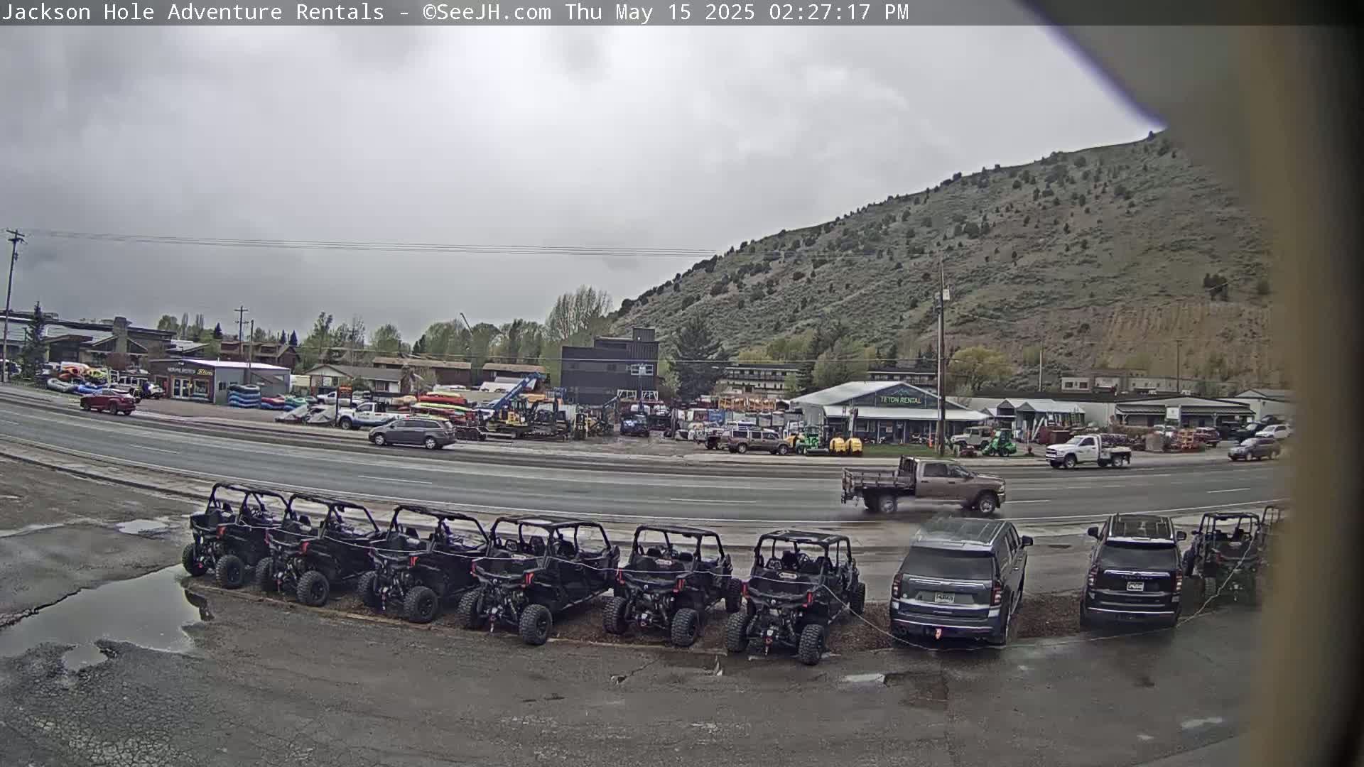 Under an overcast sky, a row of parked off-road vehicles sits in front of a rental business near a highway, with several other vehicles parked nearby and a hill visible in the background.