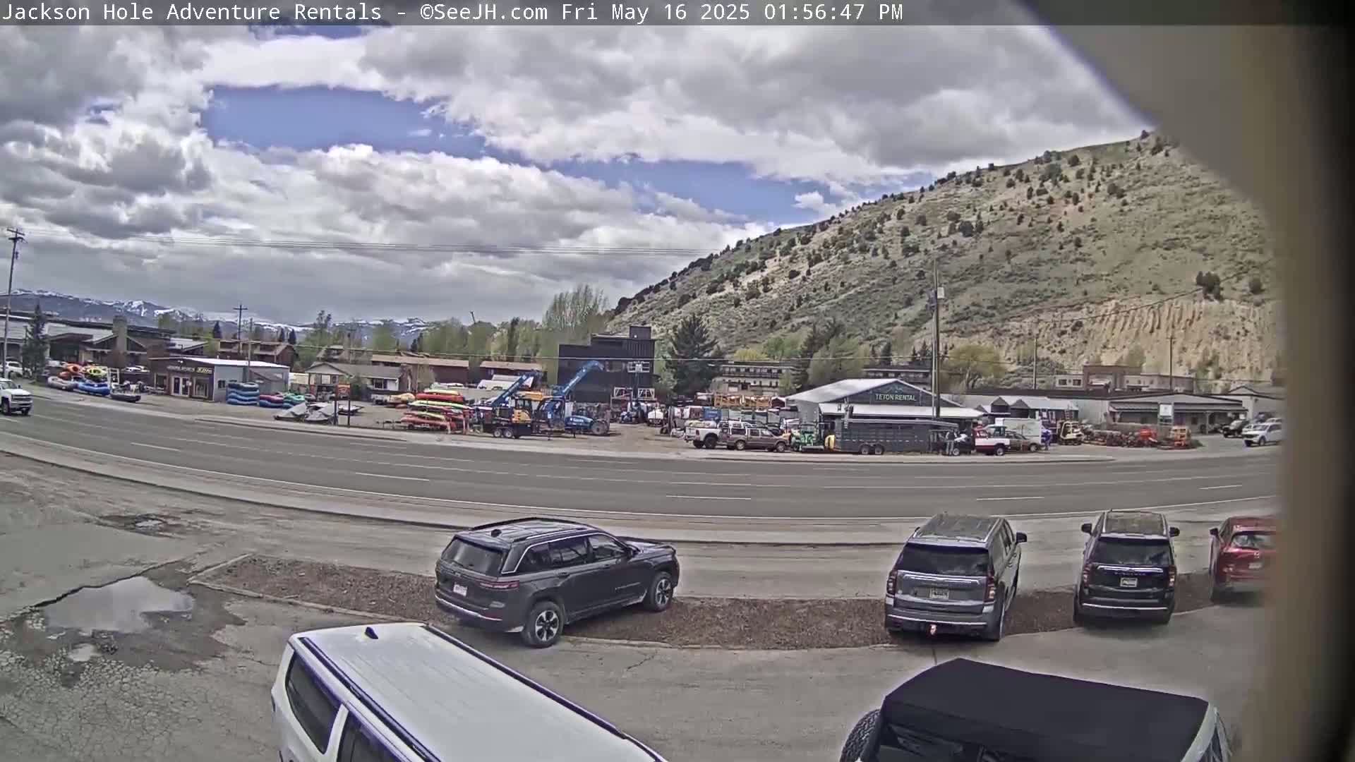A partly cloudy sky overlooks a road with several parked SUVs and a rental business with various outdoor recreational equipment, situated at the base of a tan-colored hill.