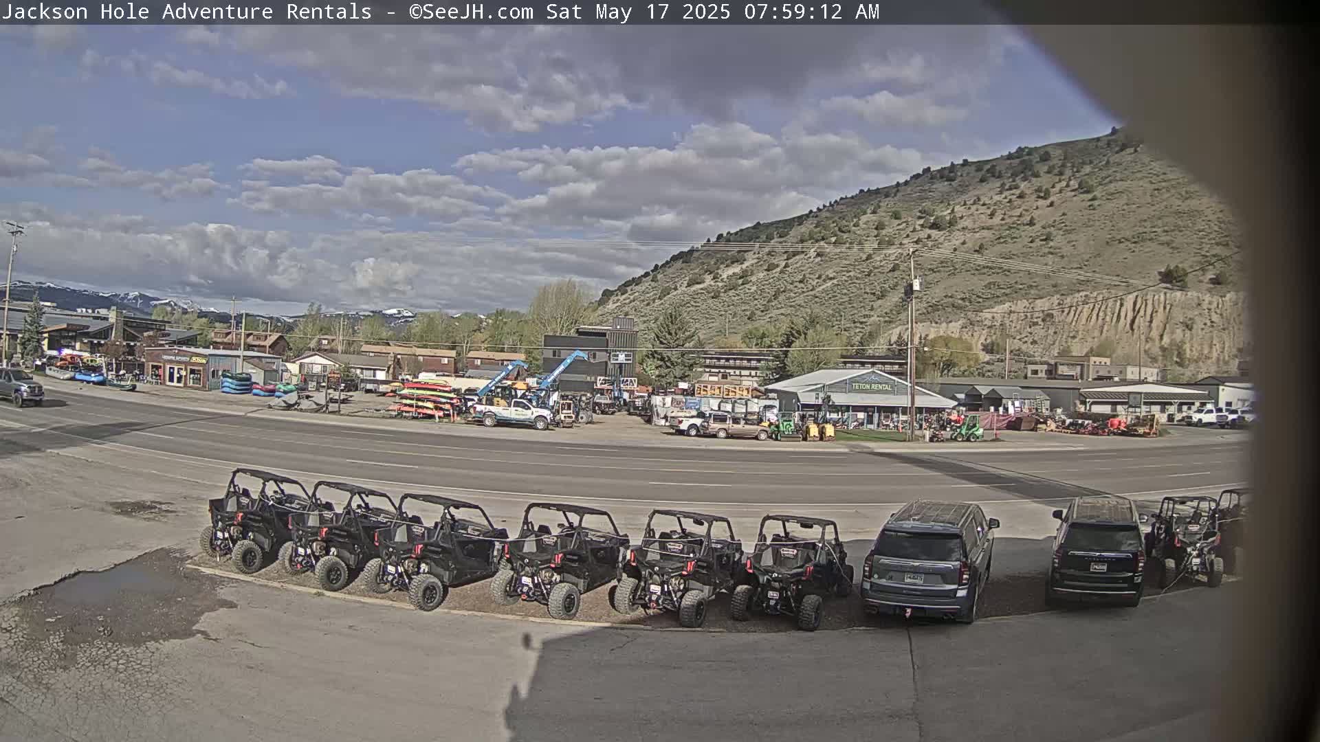 A partly cloudy day reveals a street scene with several parked ATVs and SUVs in front of a row of businesses renting outdoor recreation equipment, set against a backdrop of a dry, hilly landscape.