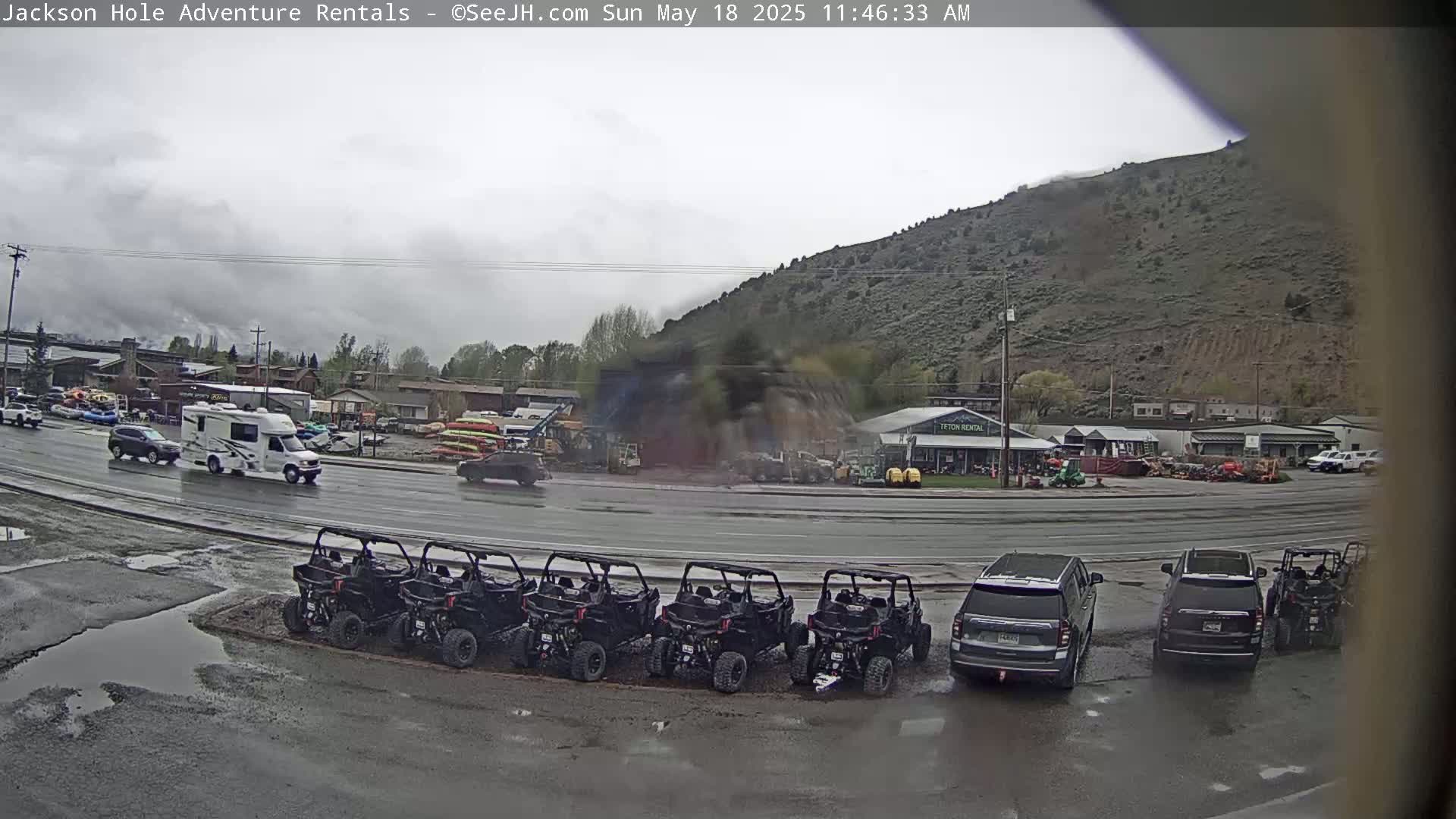 A wet street scene shows several vehicles parked, including a row of all-terrain vehicles, SUVs, and a campervan, with a hill in the background under an overcast sky.