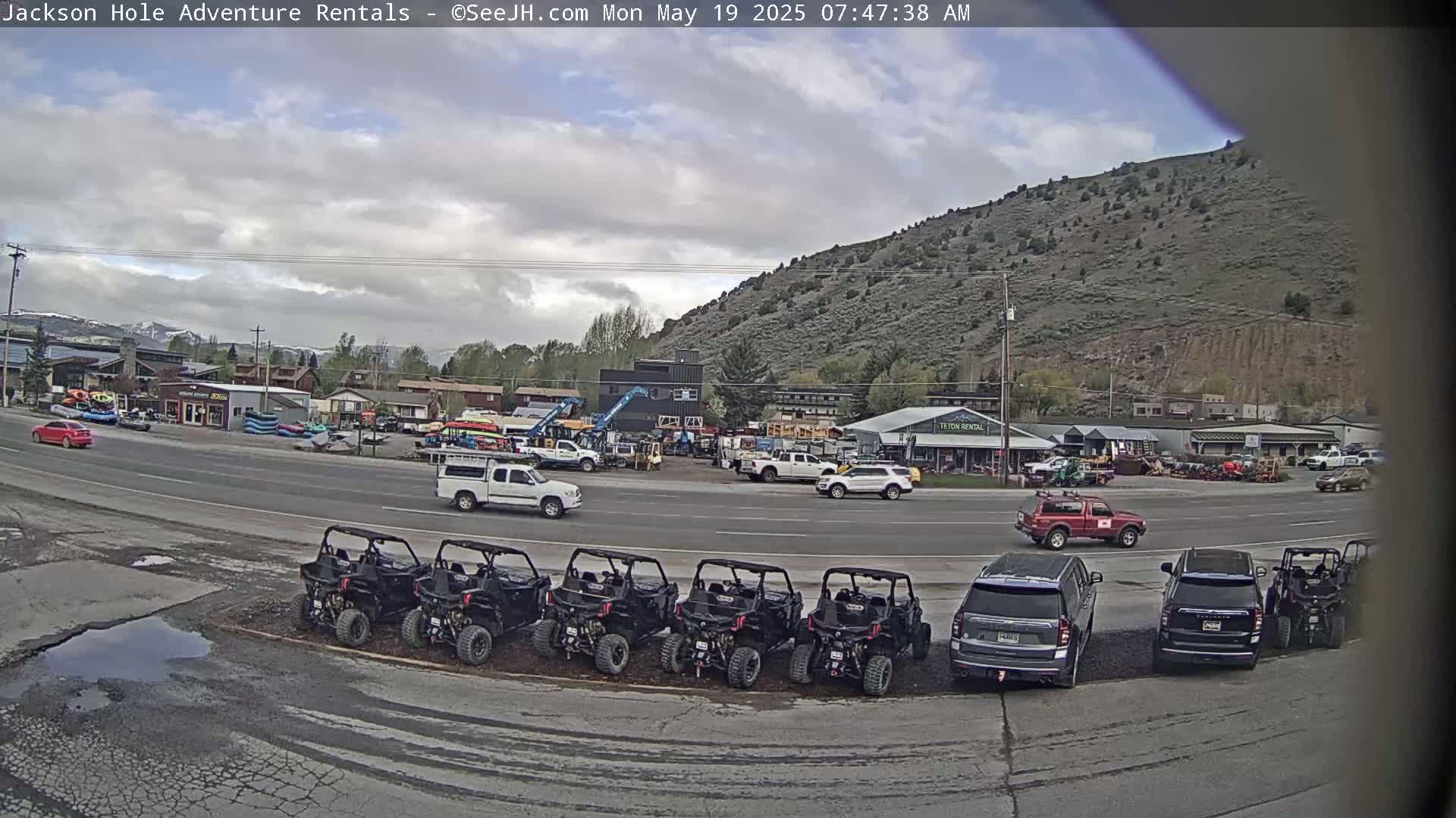 A parking lot filled with all-terrain vehicles sits in front of several businesses and a hillside, under a partly cloudy sky.