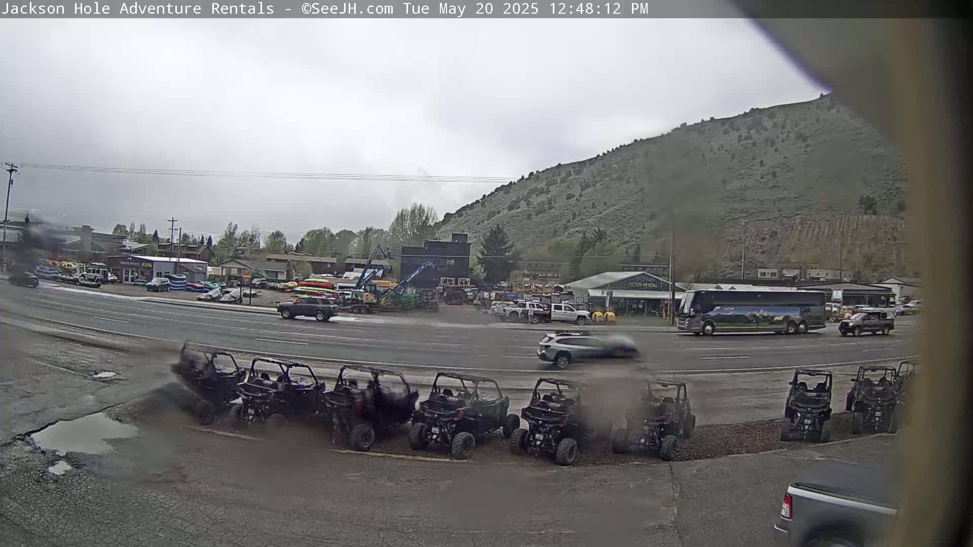 A row of all-terrain vehicles is parked in a lot near a busy road, with a large bus and several other vehicles visible, in front of a hill under an overcast sky.