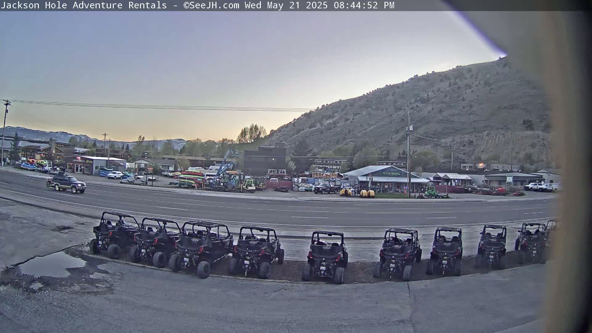 A row of off-road vehicles is parked in a lot next to a road, across from several businesses and a hillside under a twilight sky.