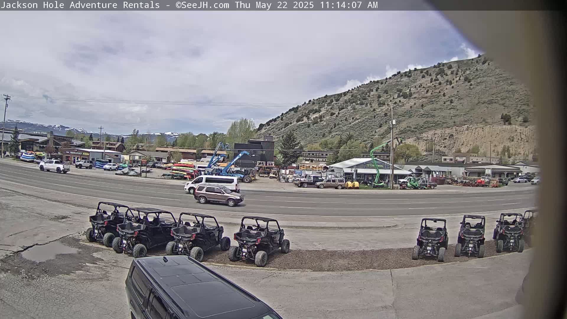 Under a partly cloudy sky, several off-road vehicles are parked in a lot next to a road lined with businesses and a mountainous backdrop.
