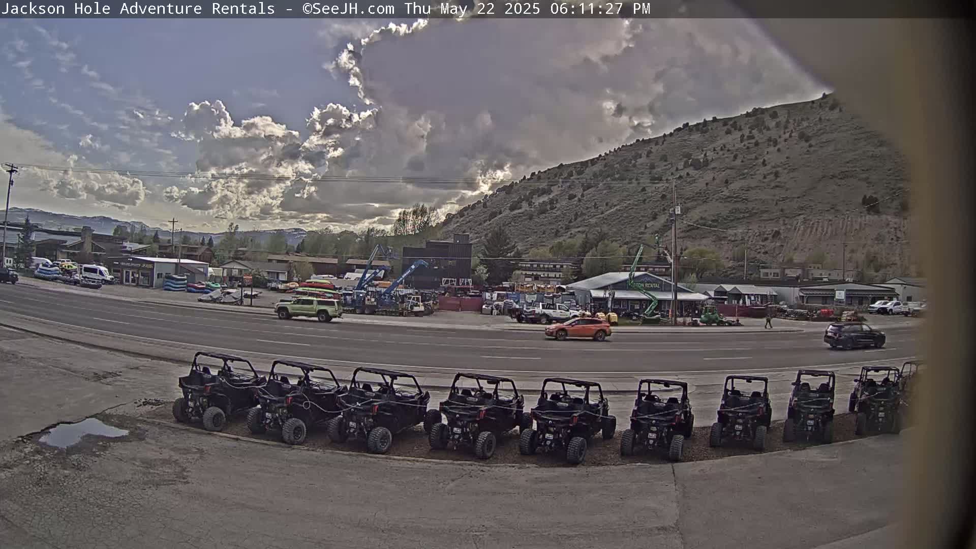 A row of all-terrain vehicles is parked in a lot near a road, with a partly cloudy sky and a mountain in the background.