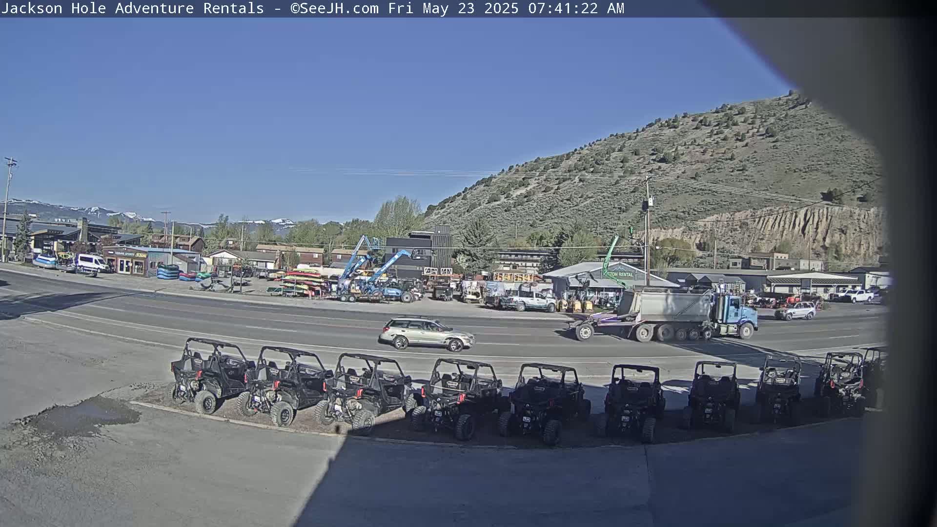 Under a clear blue sky, a row of all-terrain vehicles is parked in front of a rental business, across the street from other businesses and a large truck.