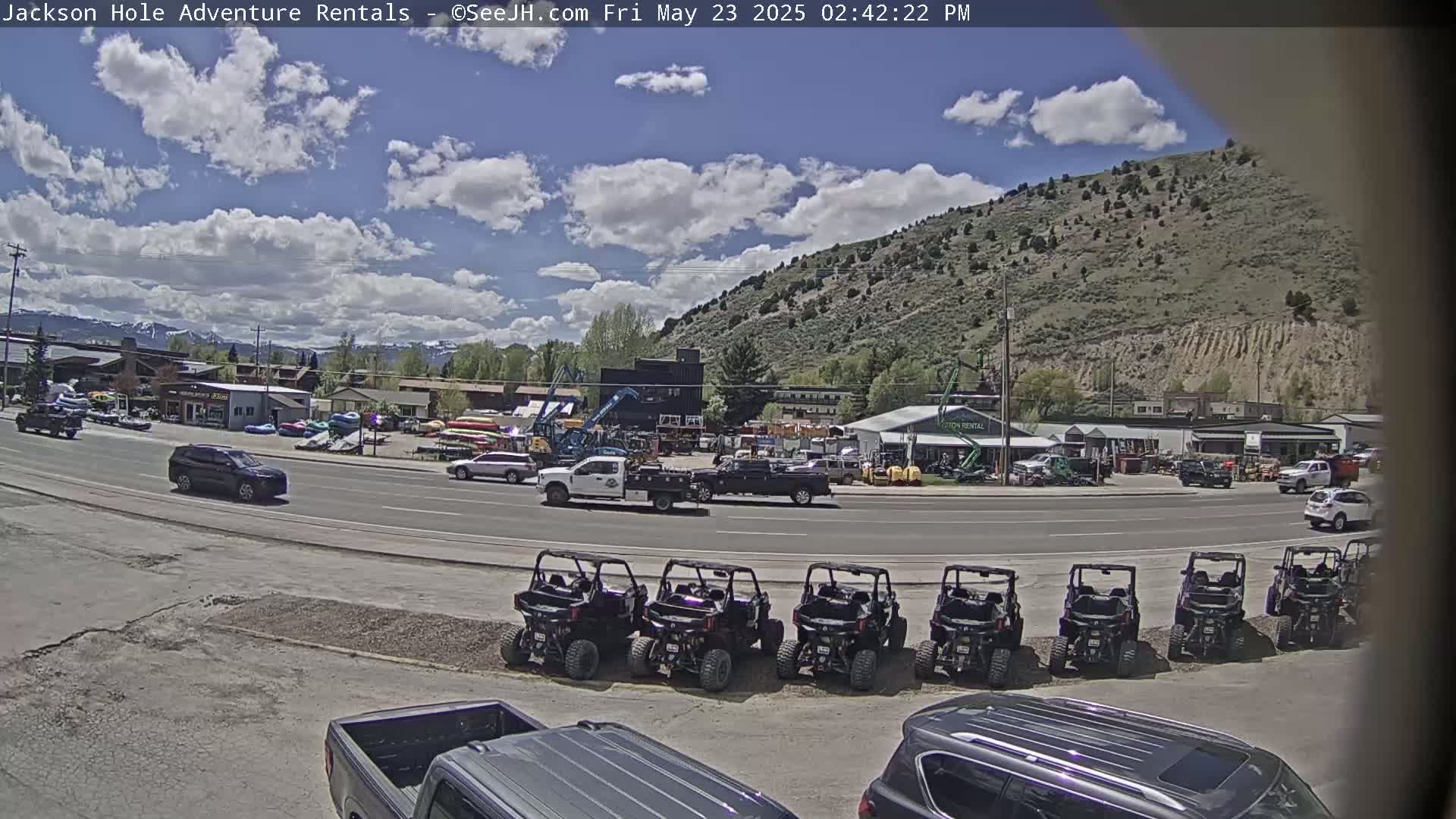 A sunny day with partly cloudy skies shows a street scene with several cars and a row of off-road vehicles parked in front of a rental business near a hillside.