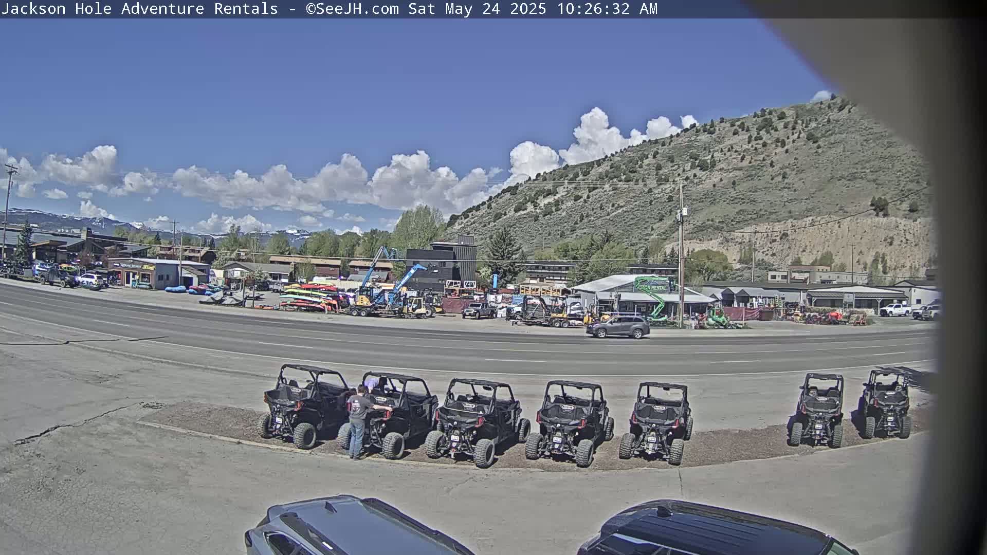 A row of off-road vehicles is parked in a lot next to a road in front of a business, under a partly cloudy sky.
