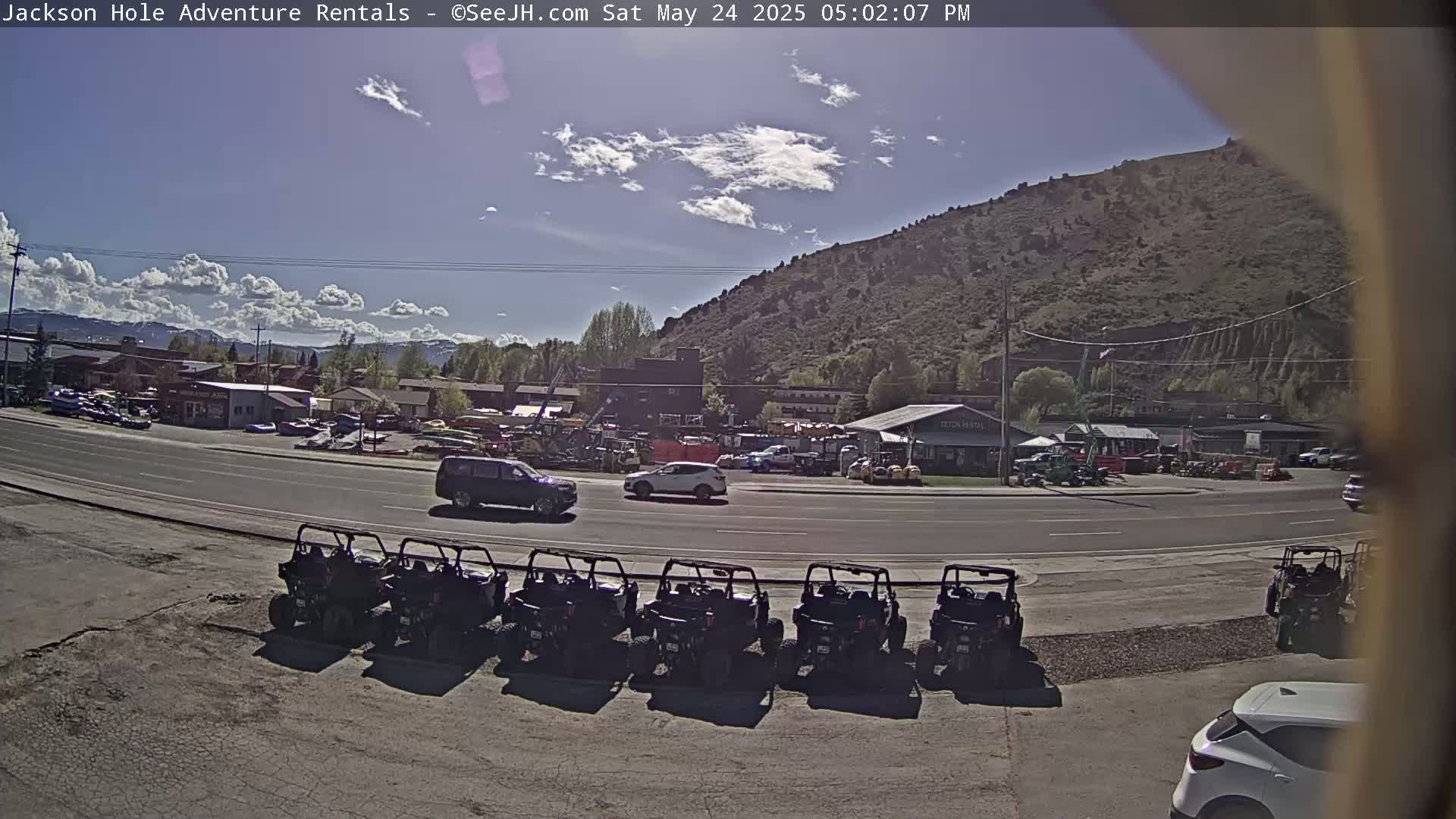 A sunny day reveals a row of parked all-terrain vehicles in front of a business, with cars driving on a road next to a hillside.