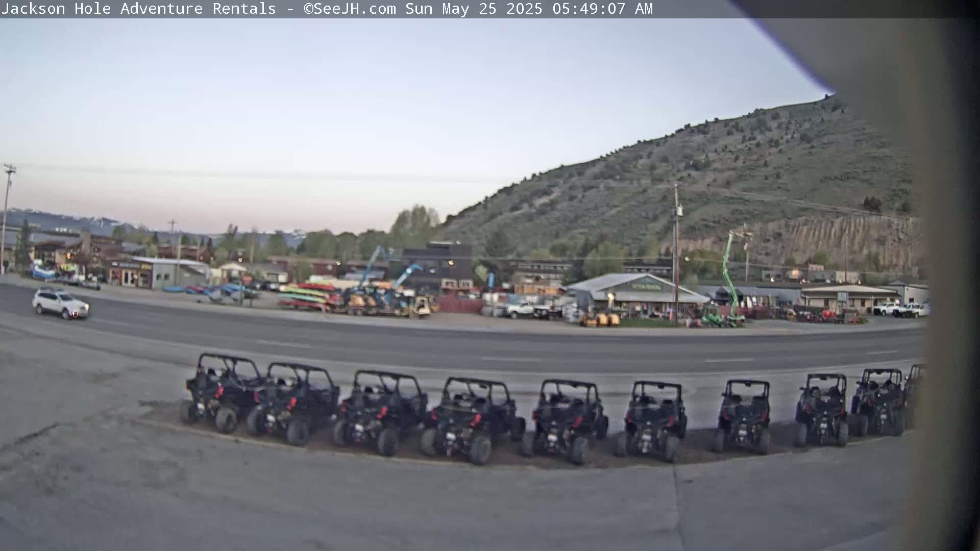 A row of all-terrain vehicles is parked in front of a building complex at the base of a hill under a clear, light-grey sky.