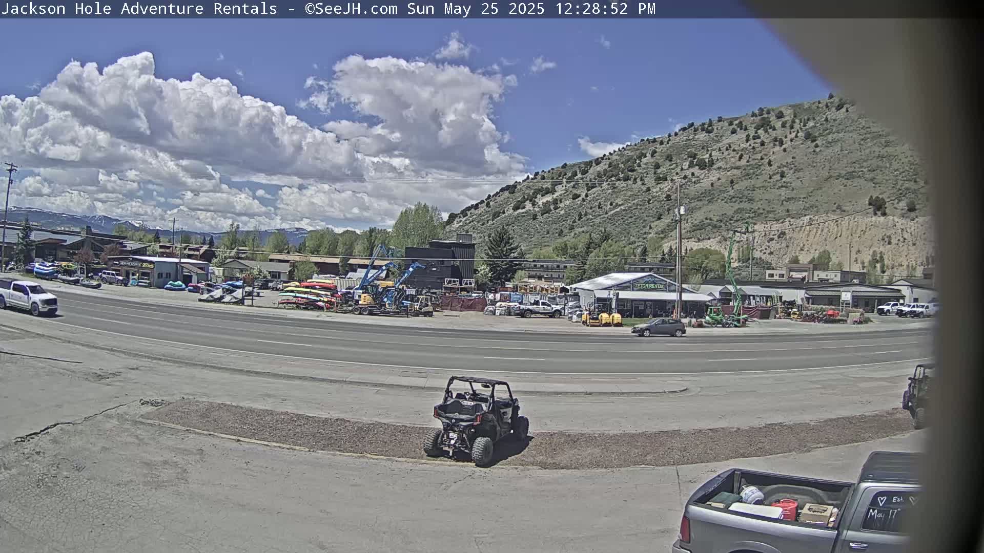 A partly cloudy day shows a street scene with various vehicles, including a four-wheeler, trucks, and construction equipment, parked near a row of businesses against a backdrop of a large, sparsely vegetated hillside.