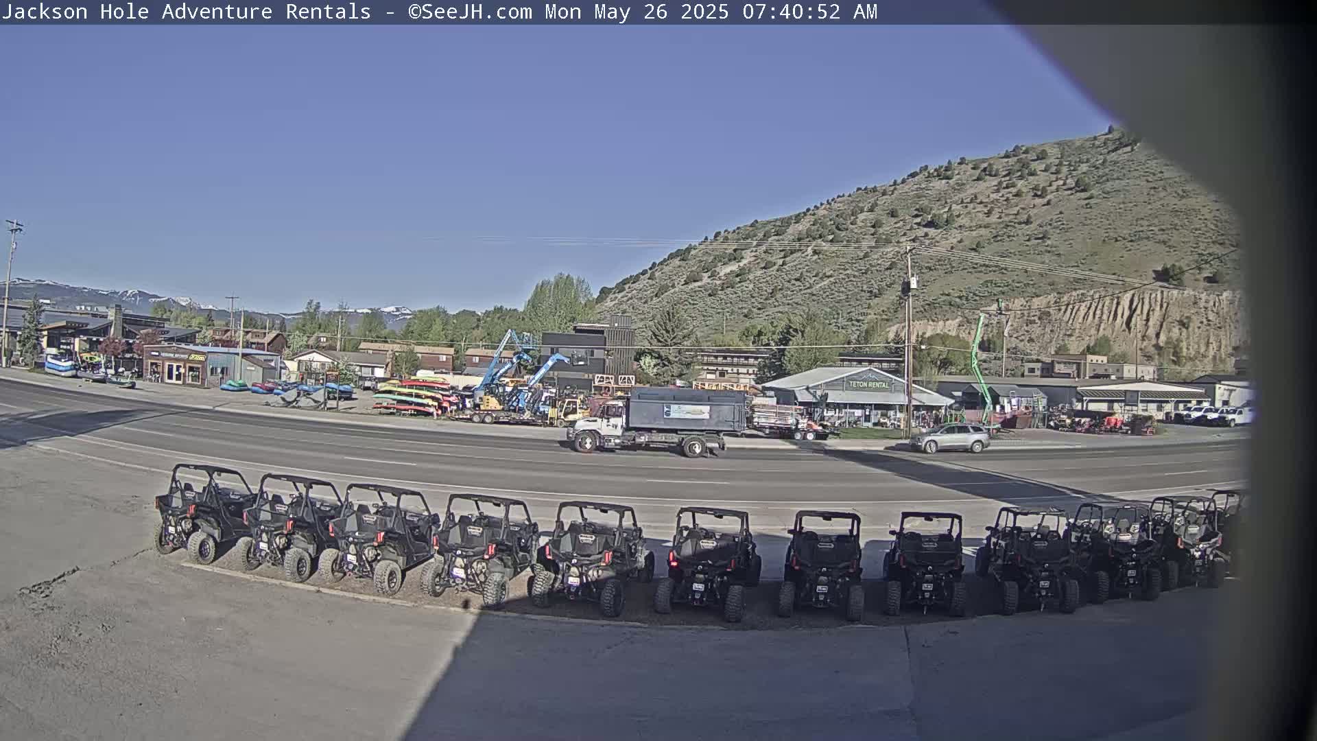 Under a clear blue sky, a row of all-terrain vehicles is parked in front of several businesses, with a truck and other vehicles nearby.