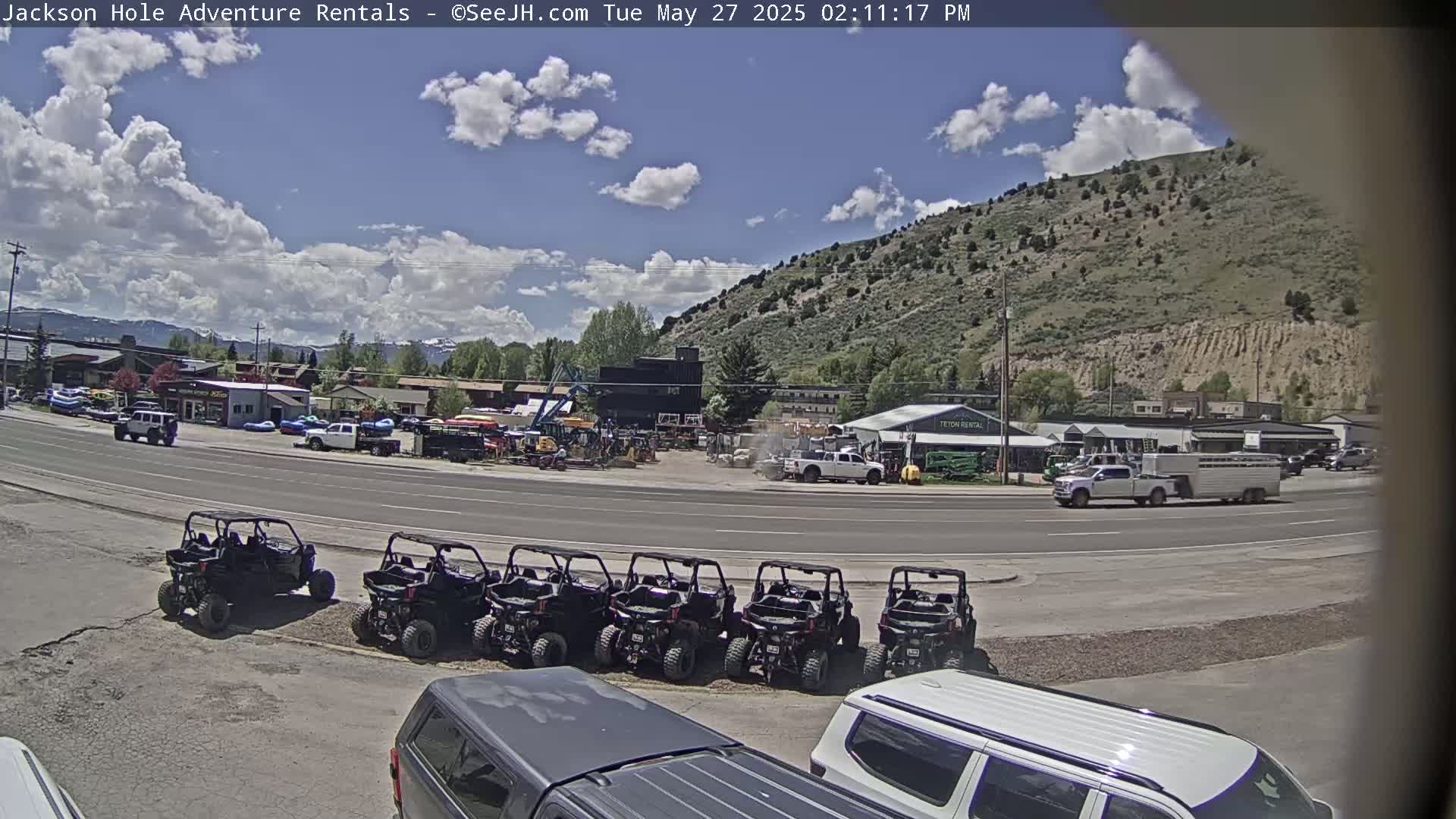 A sunny day with partly cloudy skies shows a parking lot full of off-road vehicles in front of a business complex near a mountainside.