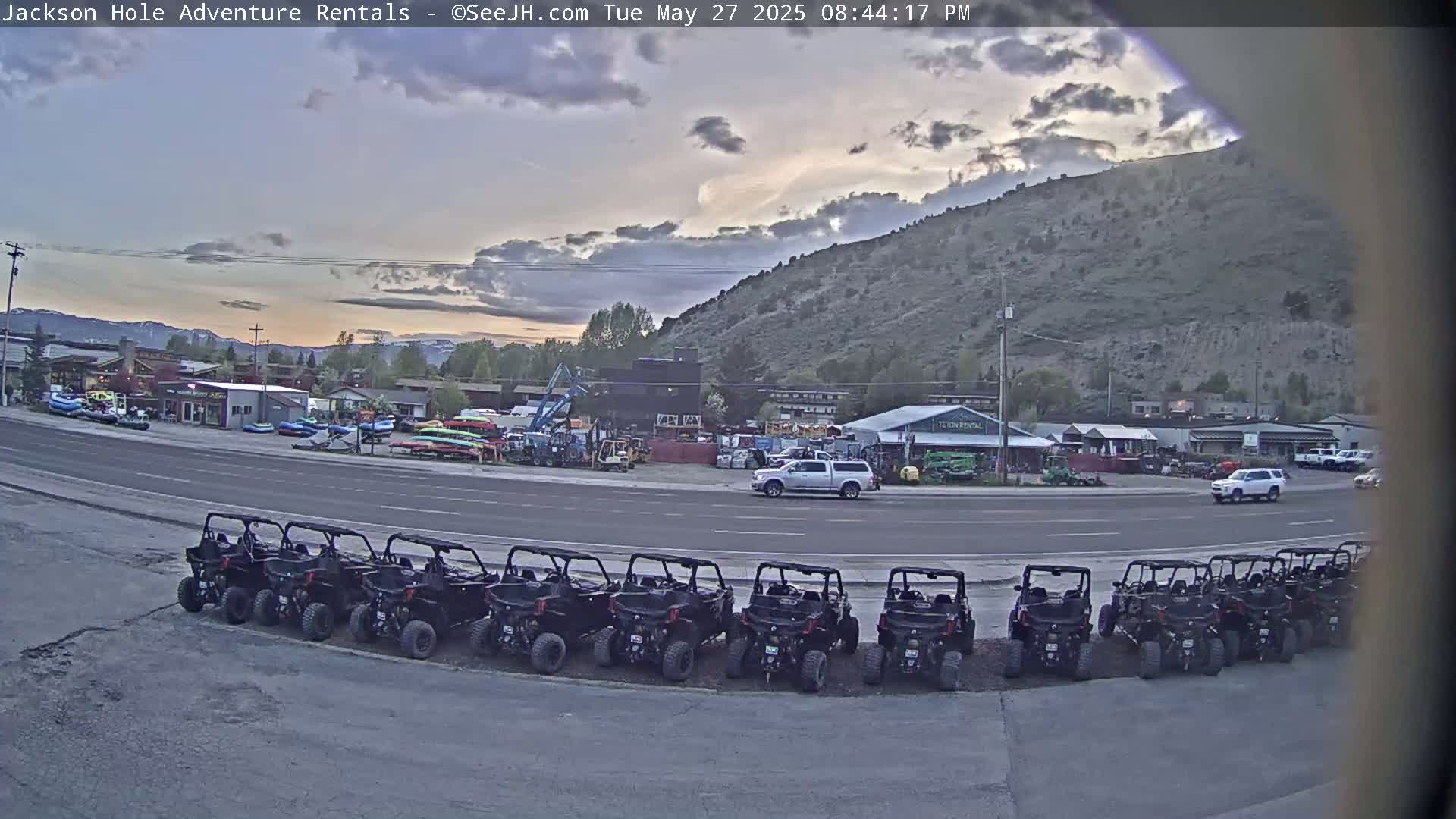 A row of all-terrain vehicles is parked in a lot next to a road, under a partly cloudy evening sky, with a mountain in the background.