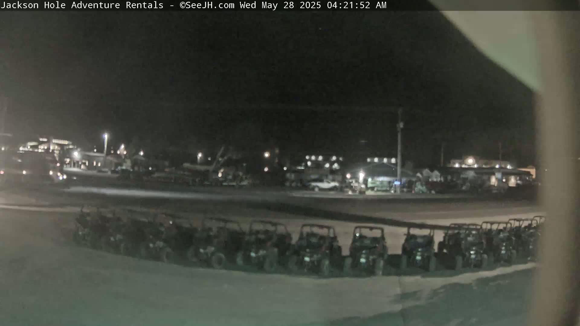 A nighttime, outdoor view of a row of parked off-road vehicles in a parking lot, with blurry buildings and lights in the background.