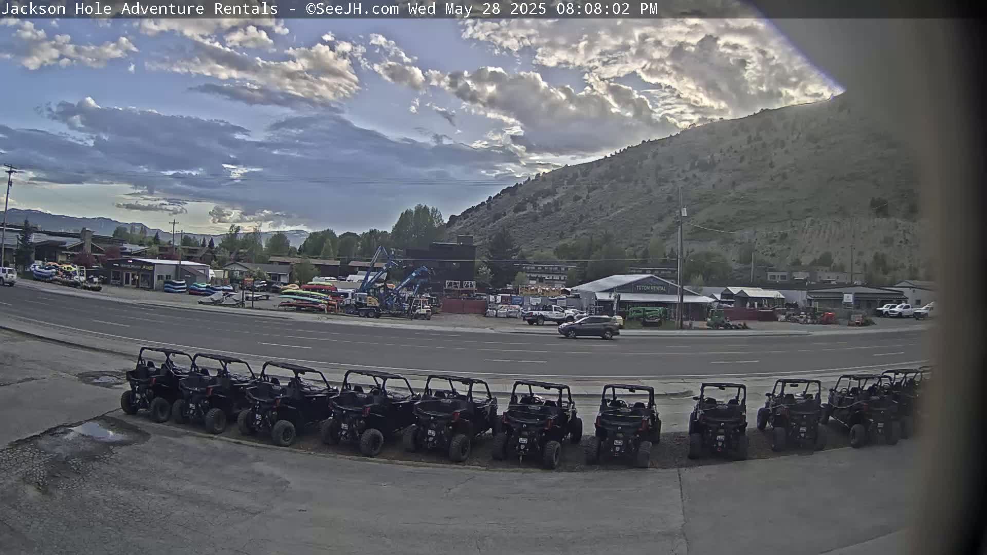 A row of off-road vehicles is parked in front of a rental business near a mountain under a partly cloudy sky.